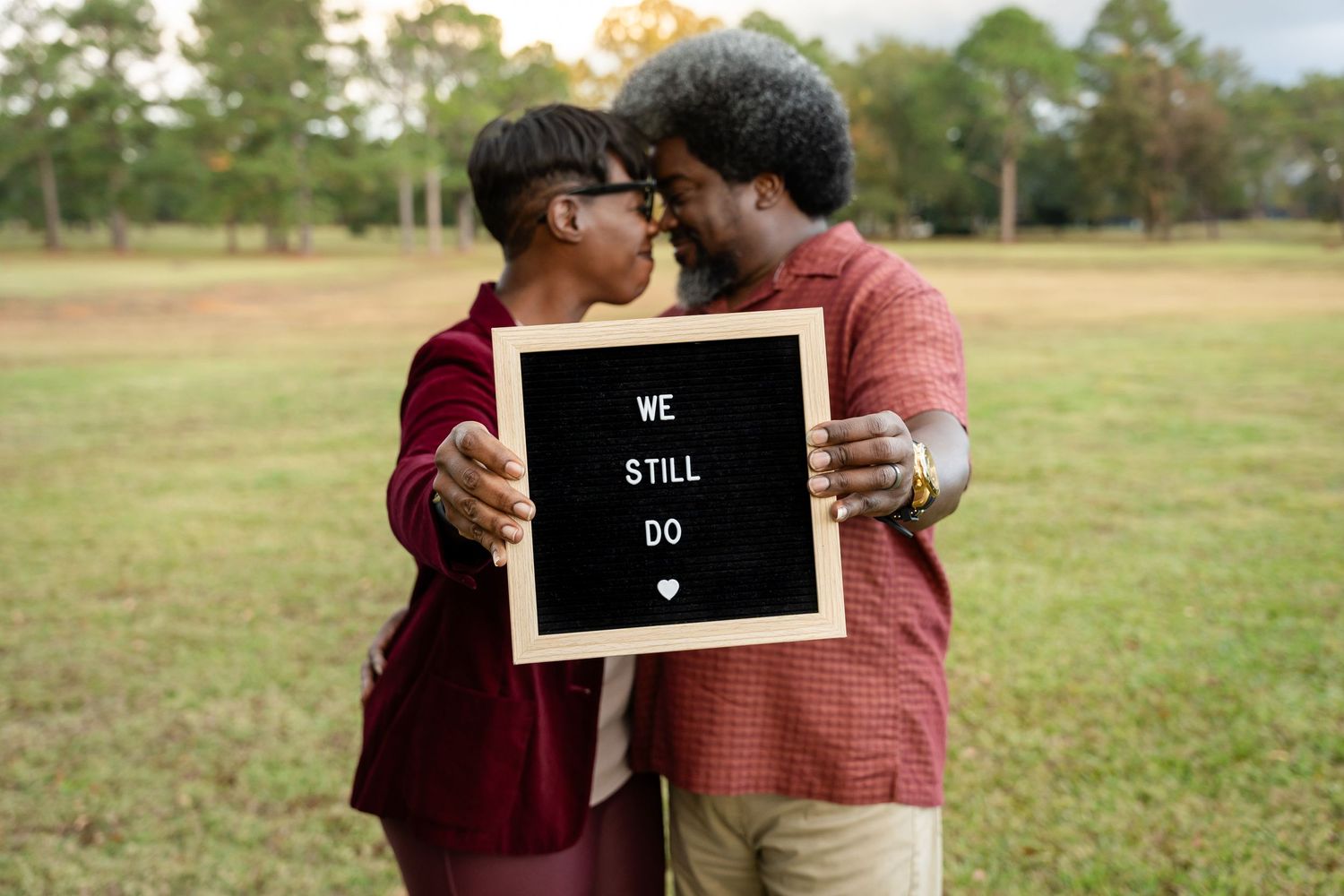 A couple poses playfully with a small chalkboard sign in a grassy field.