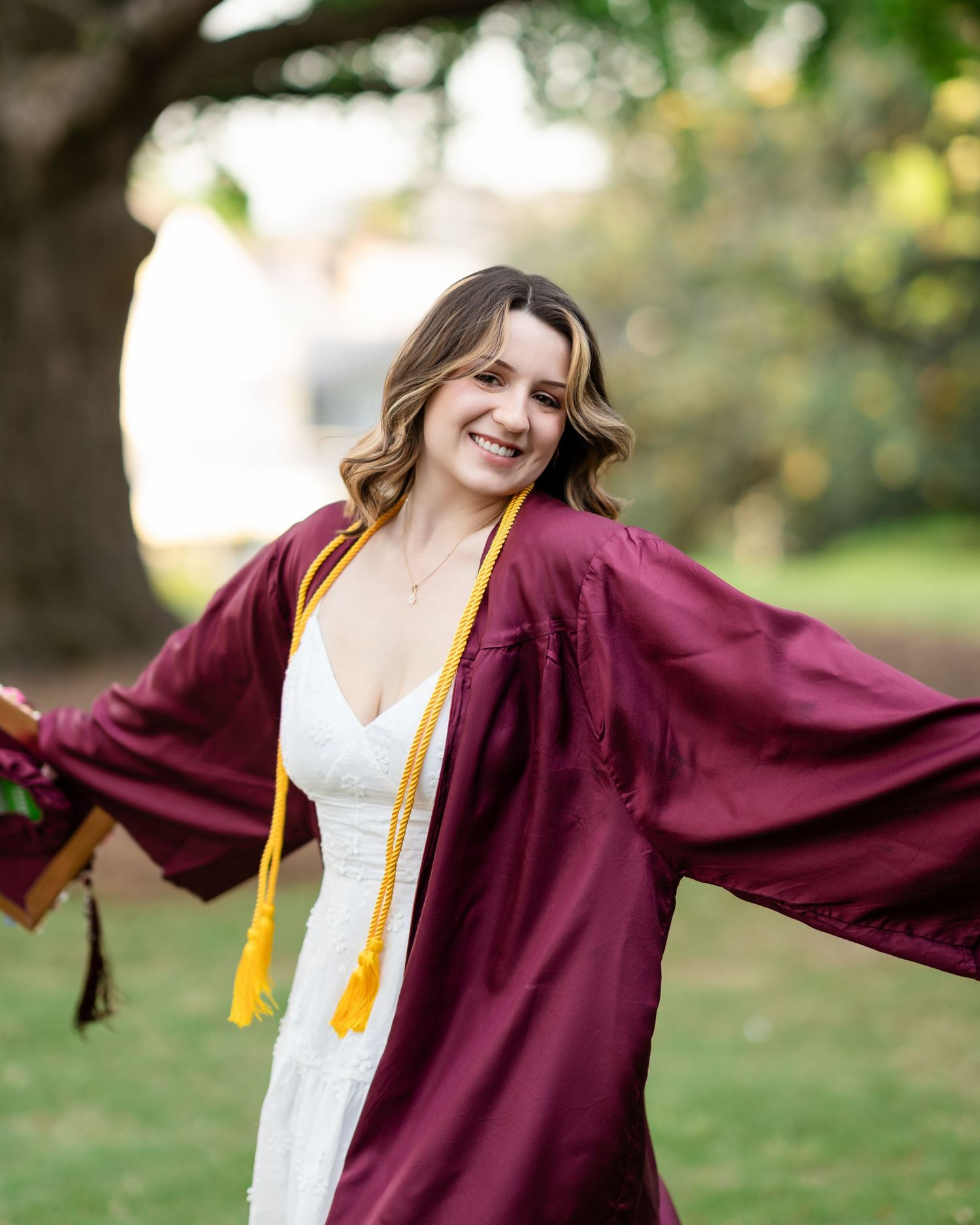 Graduate twirls in a maroon graduation gown with gold tassel against trees in natural lighting.