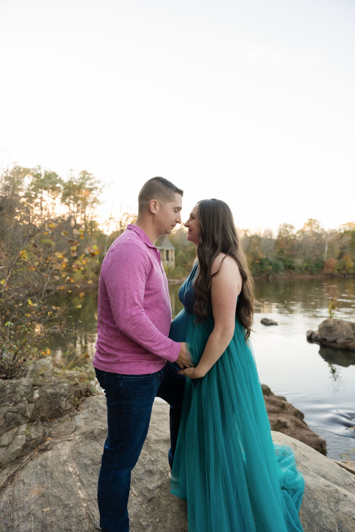 A romantic couple in formal attire embraces near a serene lake at sunset with trees in the background.
