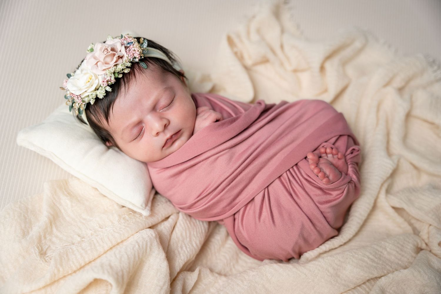 A sleeping newborn baby wrapped in dusty rose swaddle blanket lies on cream-colored fabric with floral headpiece.