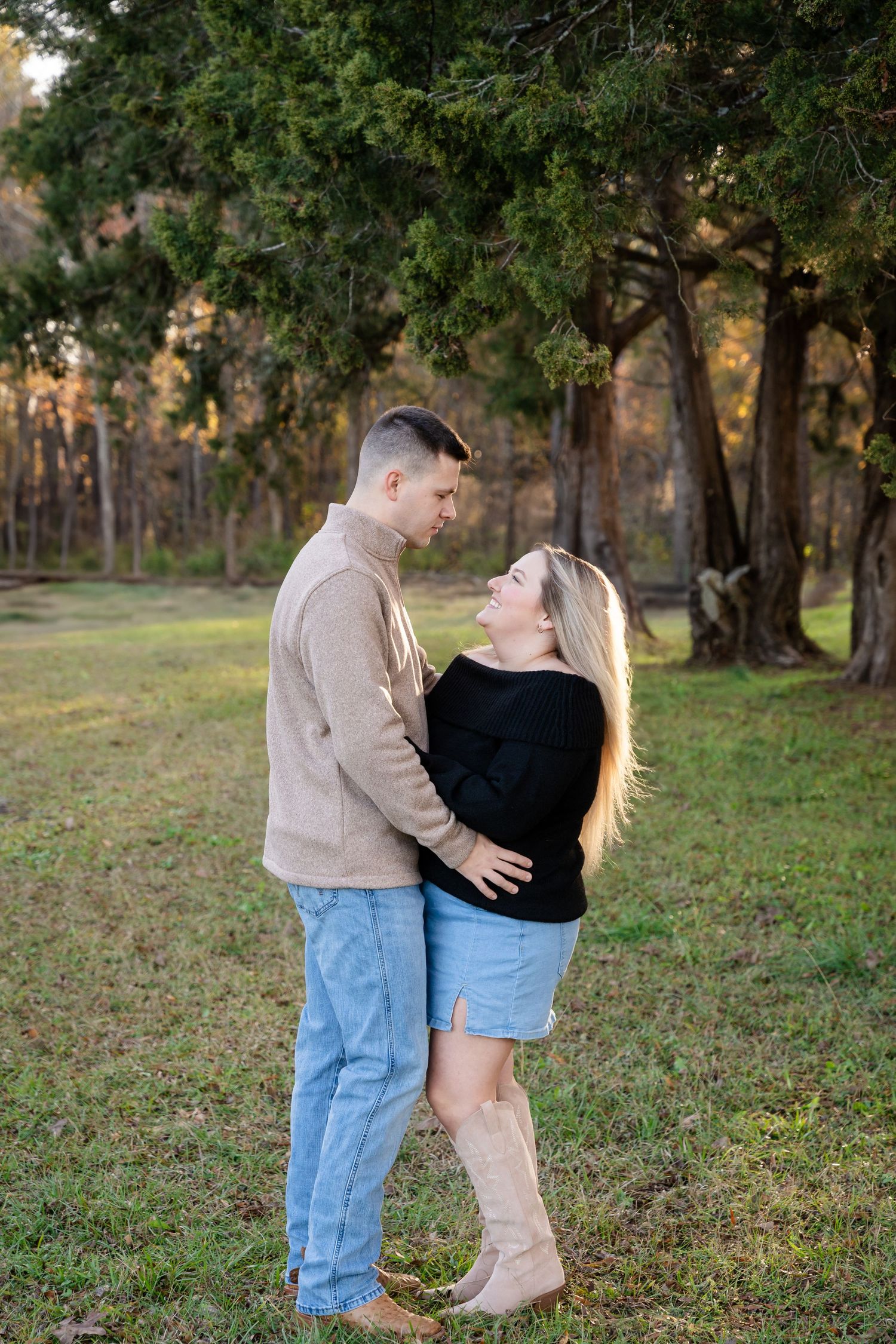 Couple embracing and smiling at each other on a grassy field surrounded by tall evergreen trees during a golden hour outdoor photo session.