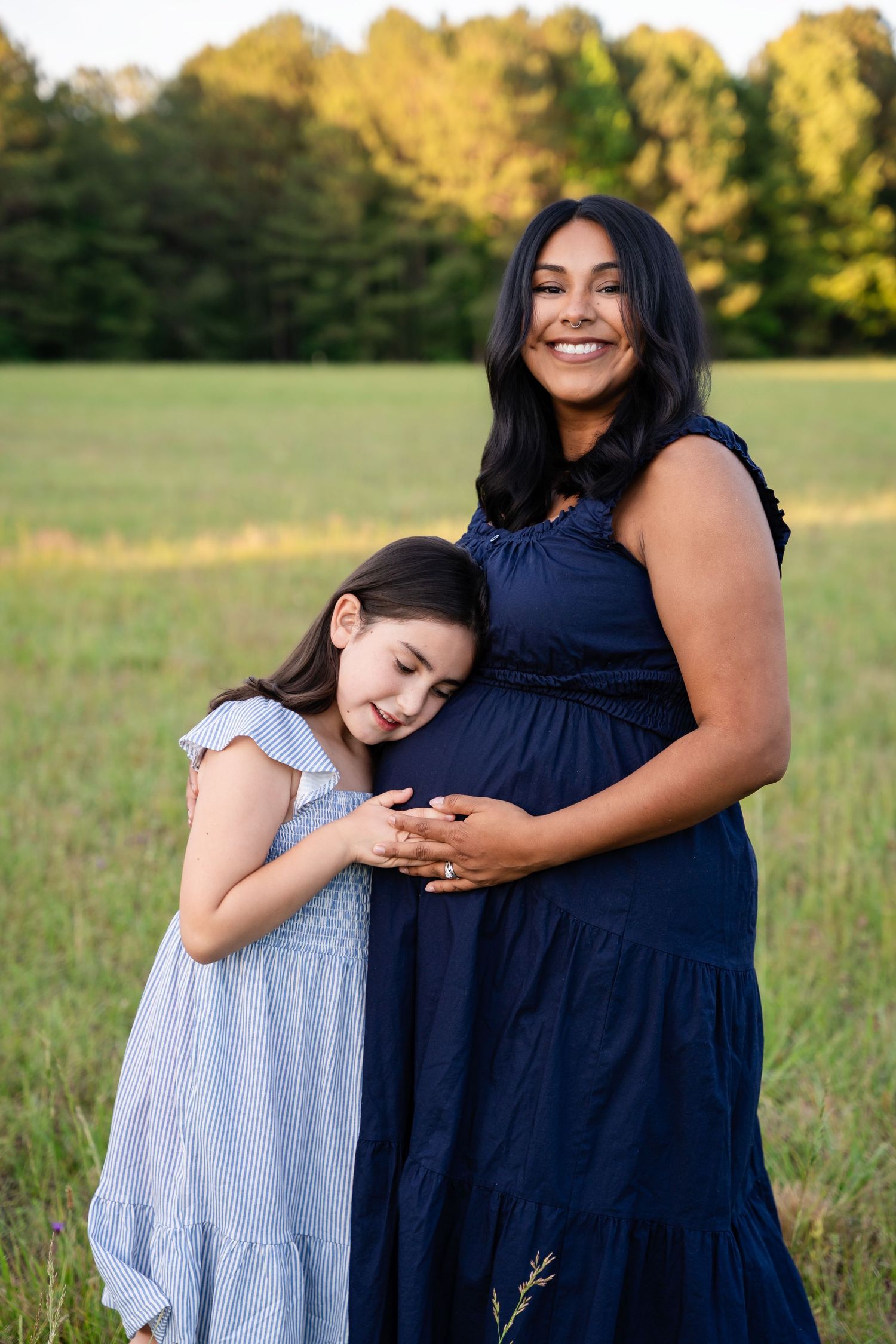 Pregnant woman in navy dress smiling as young girl hugs her baby bump in a sunny field.