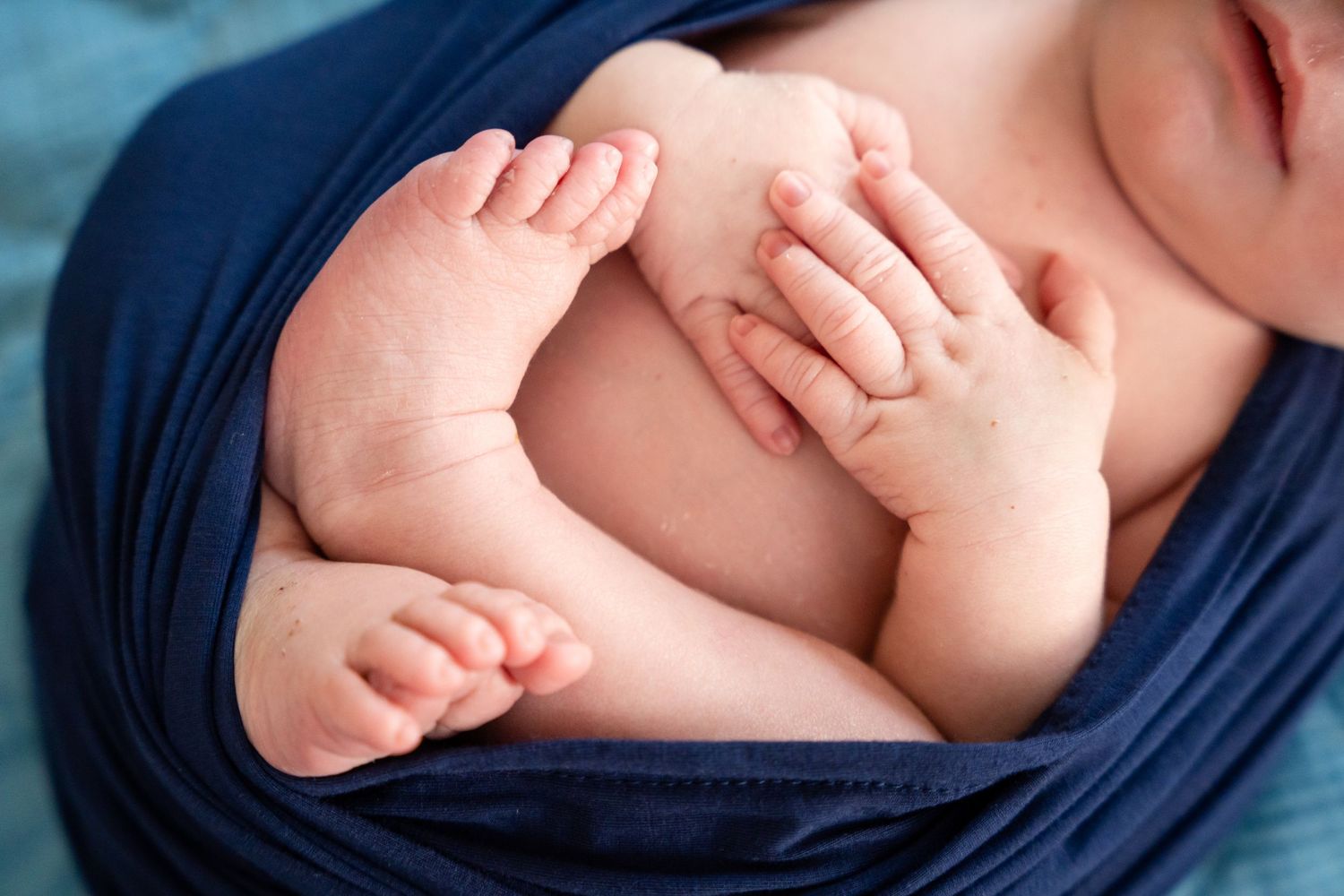 Sleeping newborn wrapped in dark blue fabric with tiny hands visible.