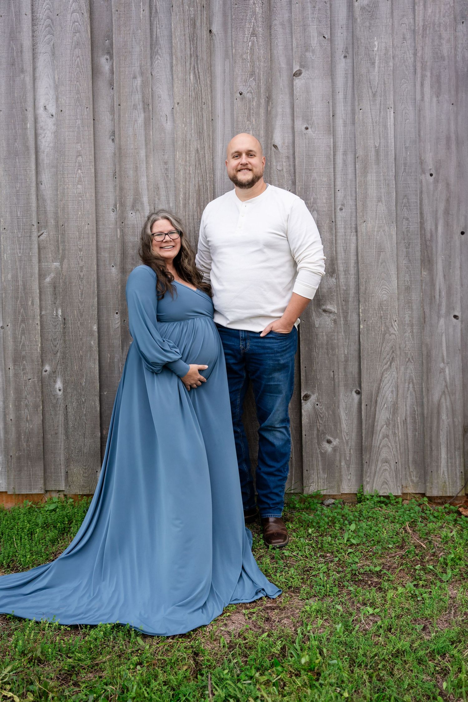 Couple poses casually in front of rustic wooden fence during outdoor photo session.