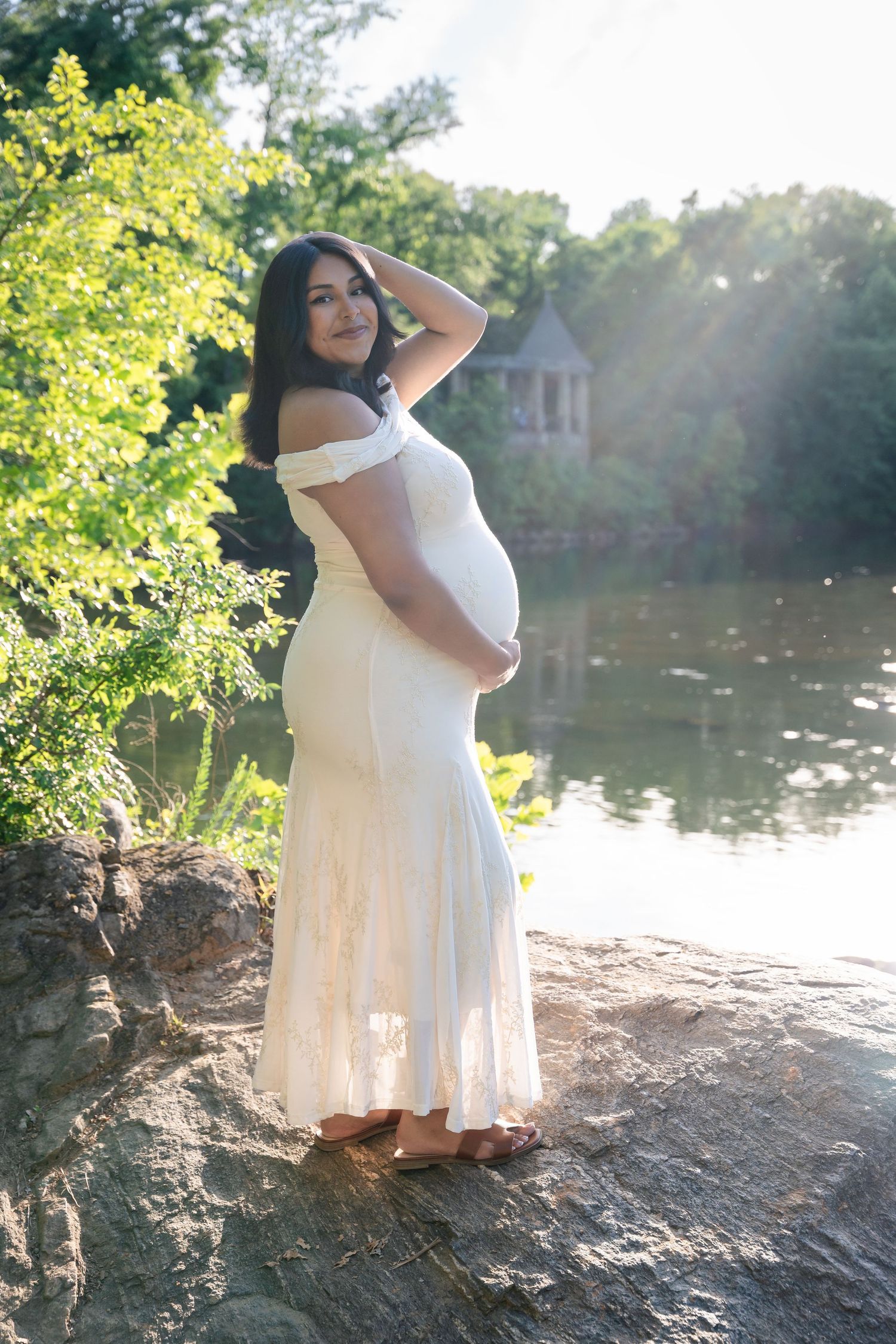 Pregnant woman in a white off-the-shoulder gown poses on a rock by a sunlit lake, with a gazebo and green trees behind her.