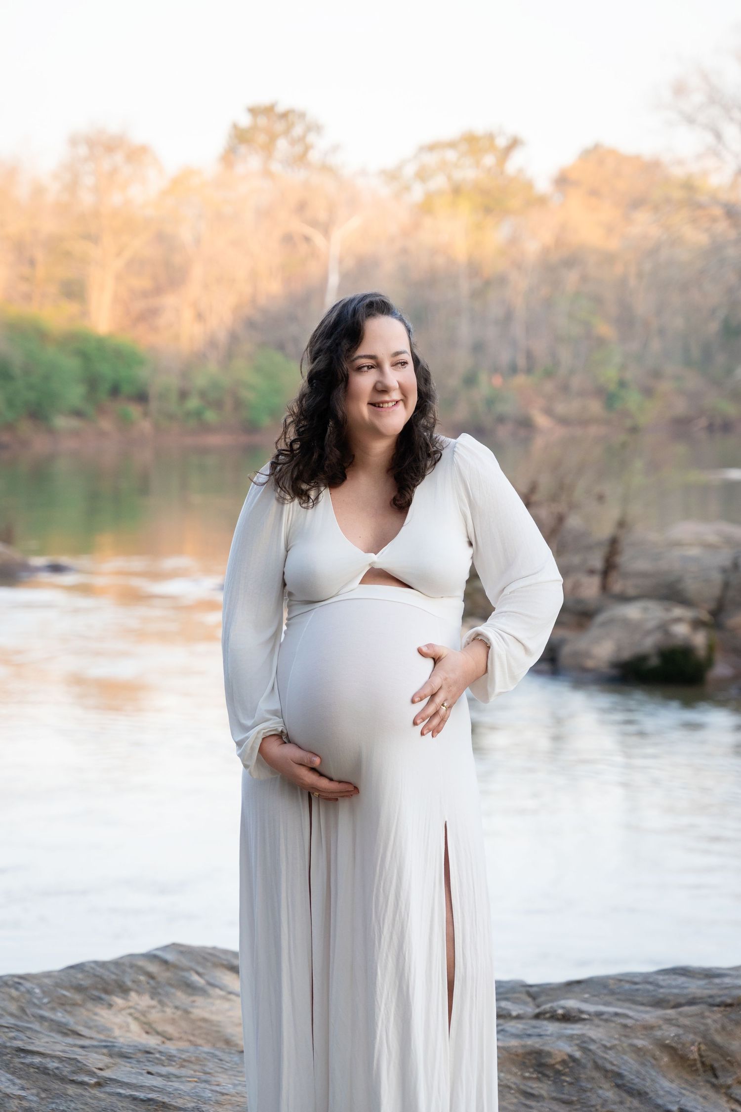 A pregnant woman wearing a long white dress stands by a river surrounded by trees in the background.