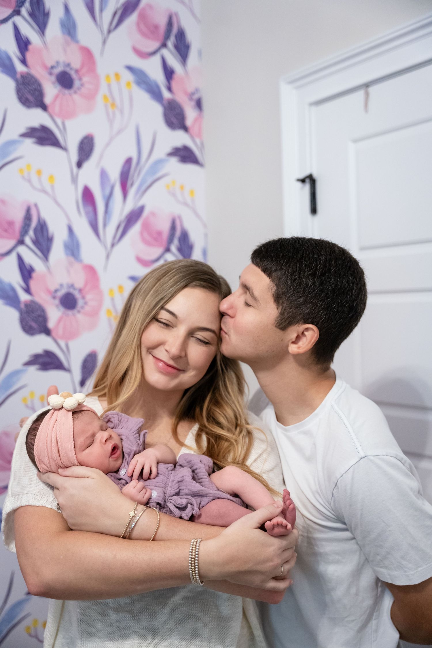 New parents share a tender moment, father kissing mother's cheek as she holds their newborn in purple outfit.