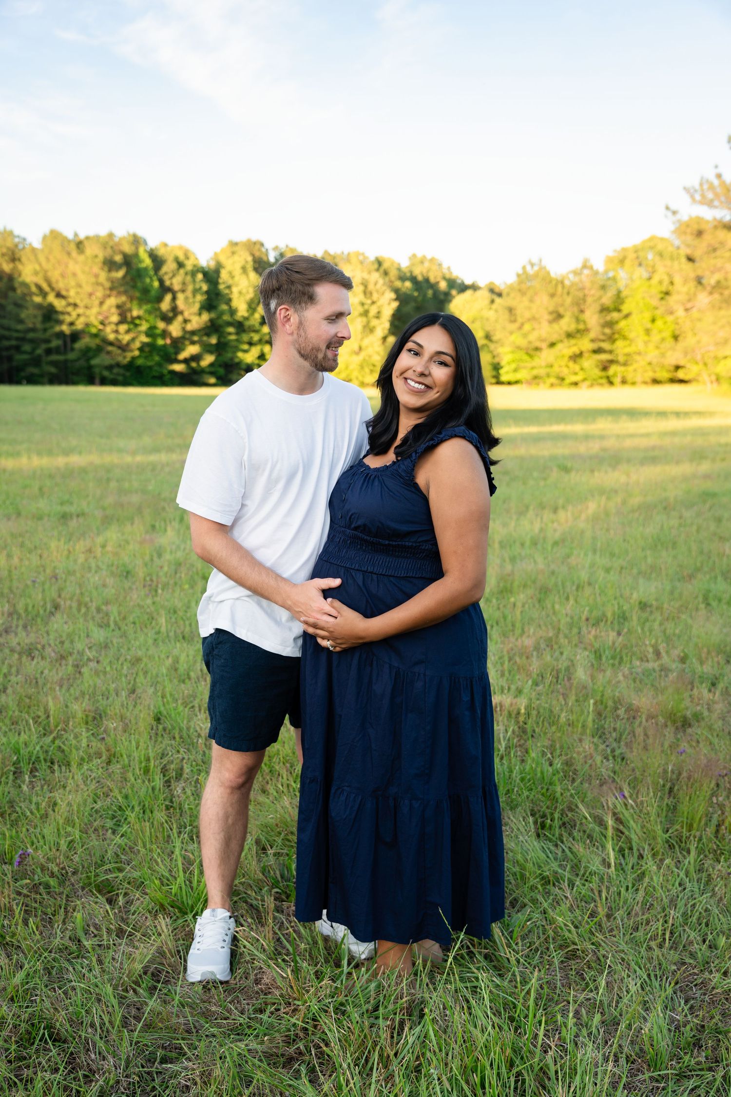 Pregnant couple embracing in a green field at golden hour, woman in navy dress cradling baby bump, man in white tee smiling.