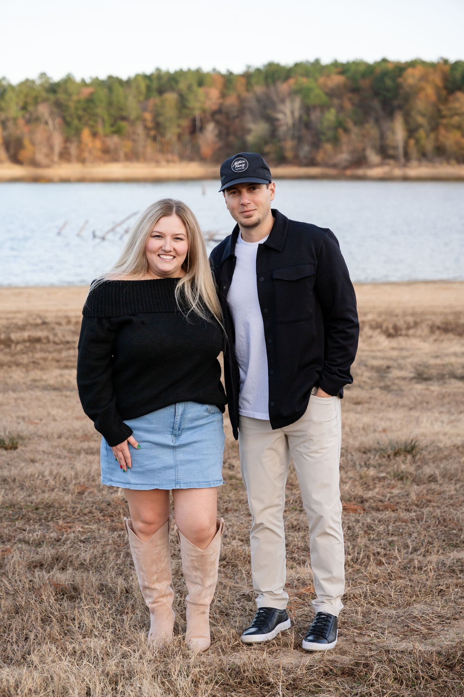A couple poses together on a sandy beach near a lake during sunset.