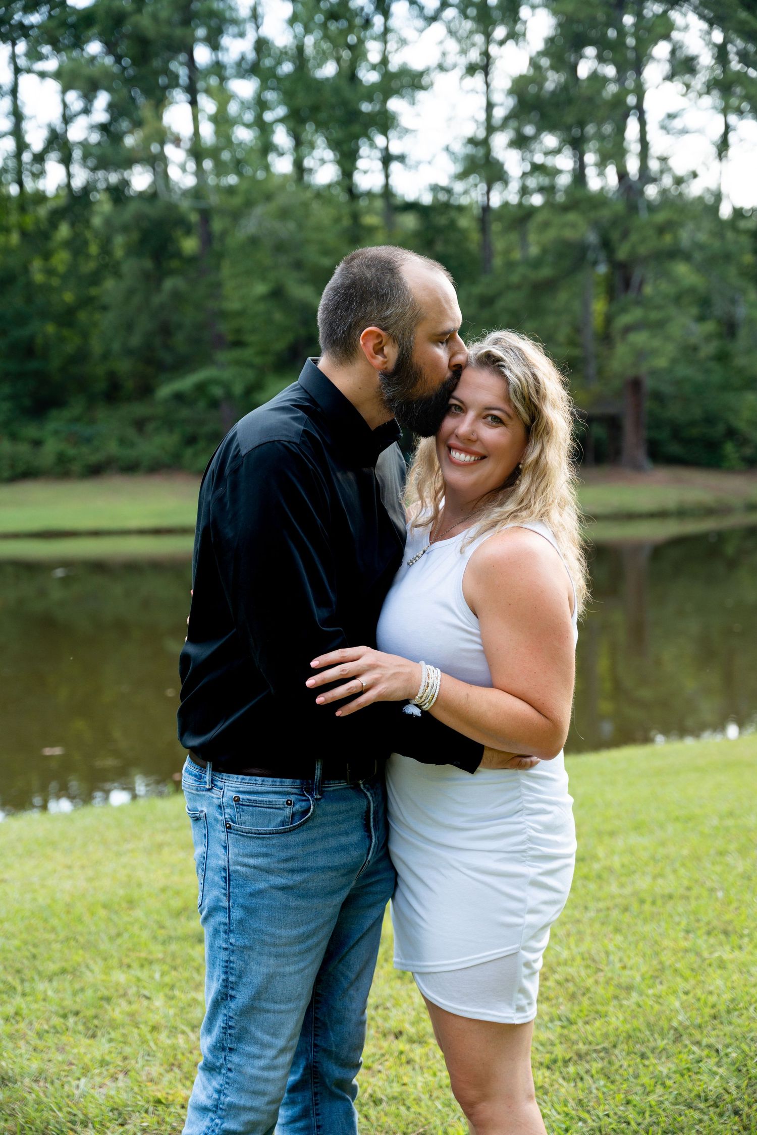 Romantic couple embracing on grass near lake with forest backdrop.