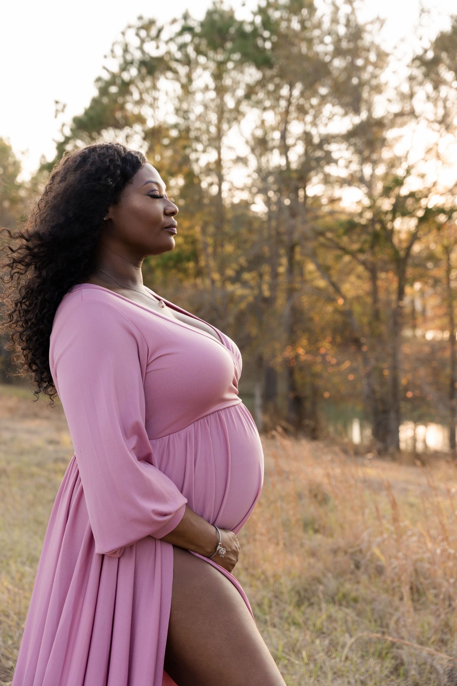 Pregnant Black woman in pink maternity dress poses outdoors in autumn field at golden hour.