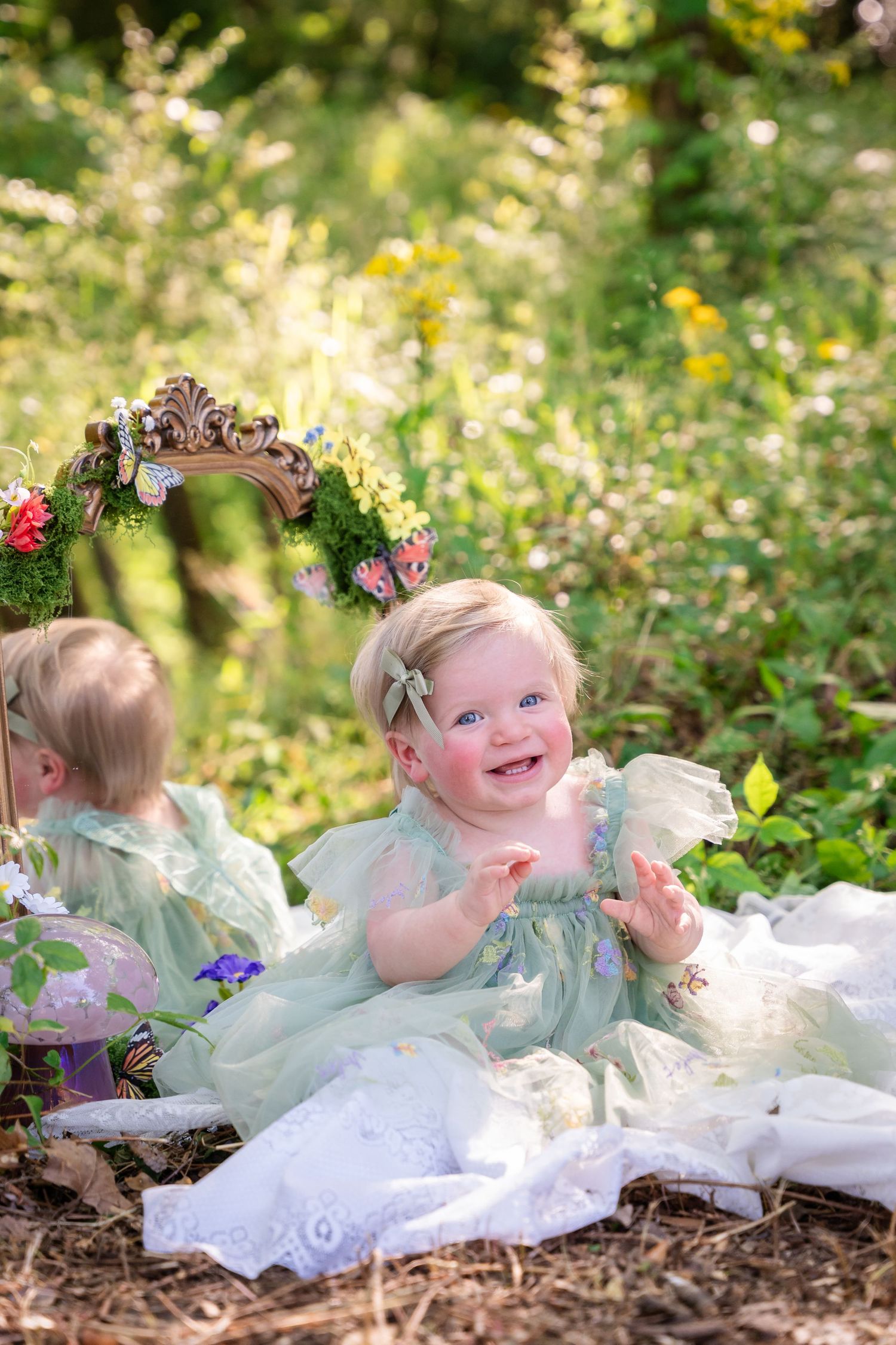 A smiling baby girl in a mint green dress sits outdoors near a ornate mirror surrounded by lush greenery.