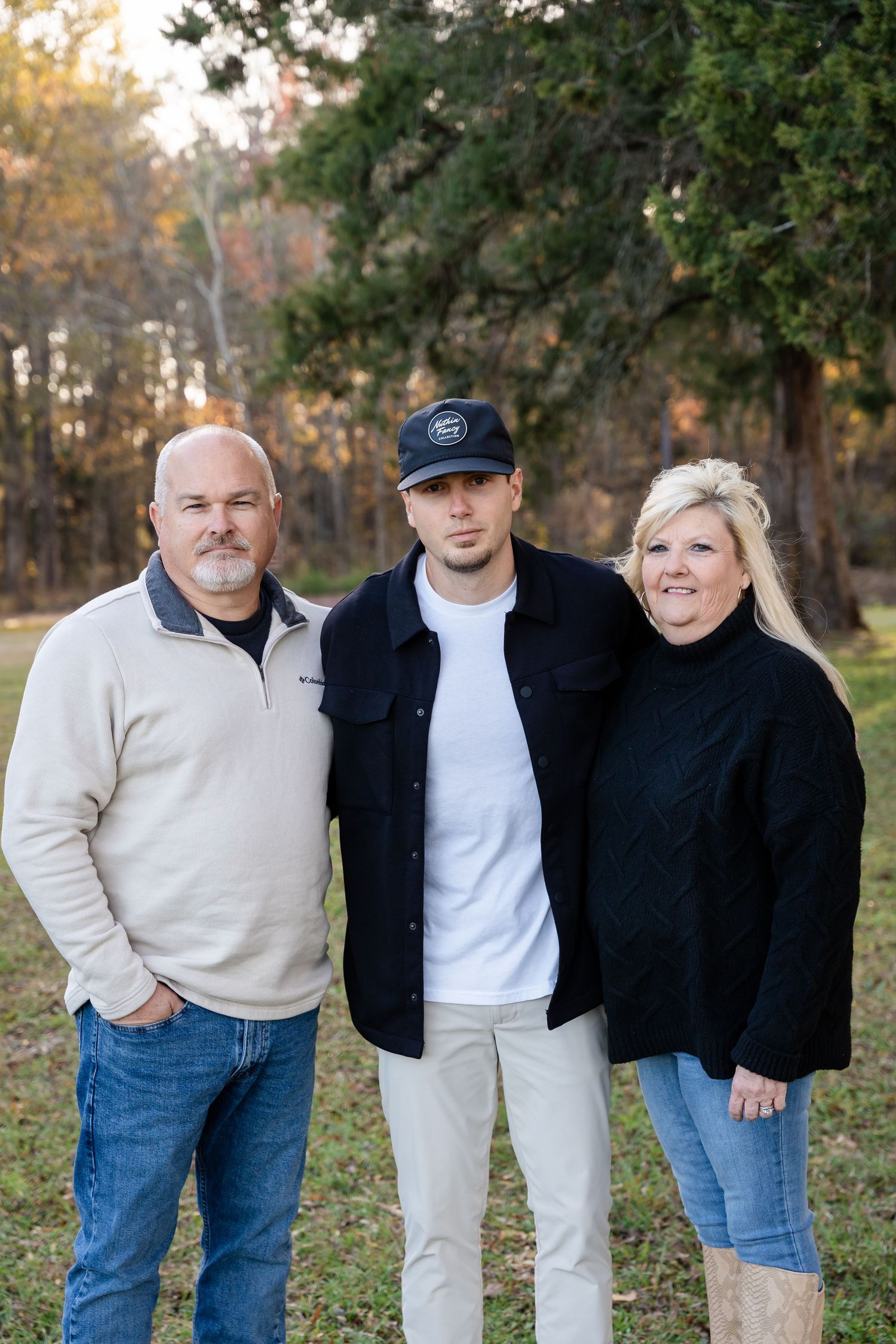 Three people pose together outdoors in a wooded park, wearing casual fall clothing, with trees and soft sunlight in the background.