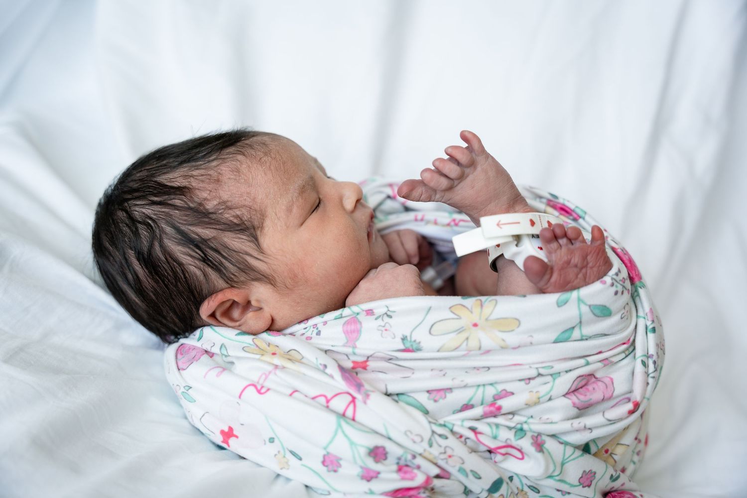 Newborn baby with dark hair sleeping peacefully on a white bed, swaddled in a soft floral blanket with a hospital ID bracelet on the wrist.