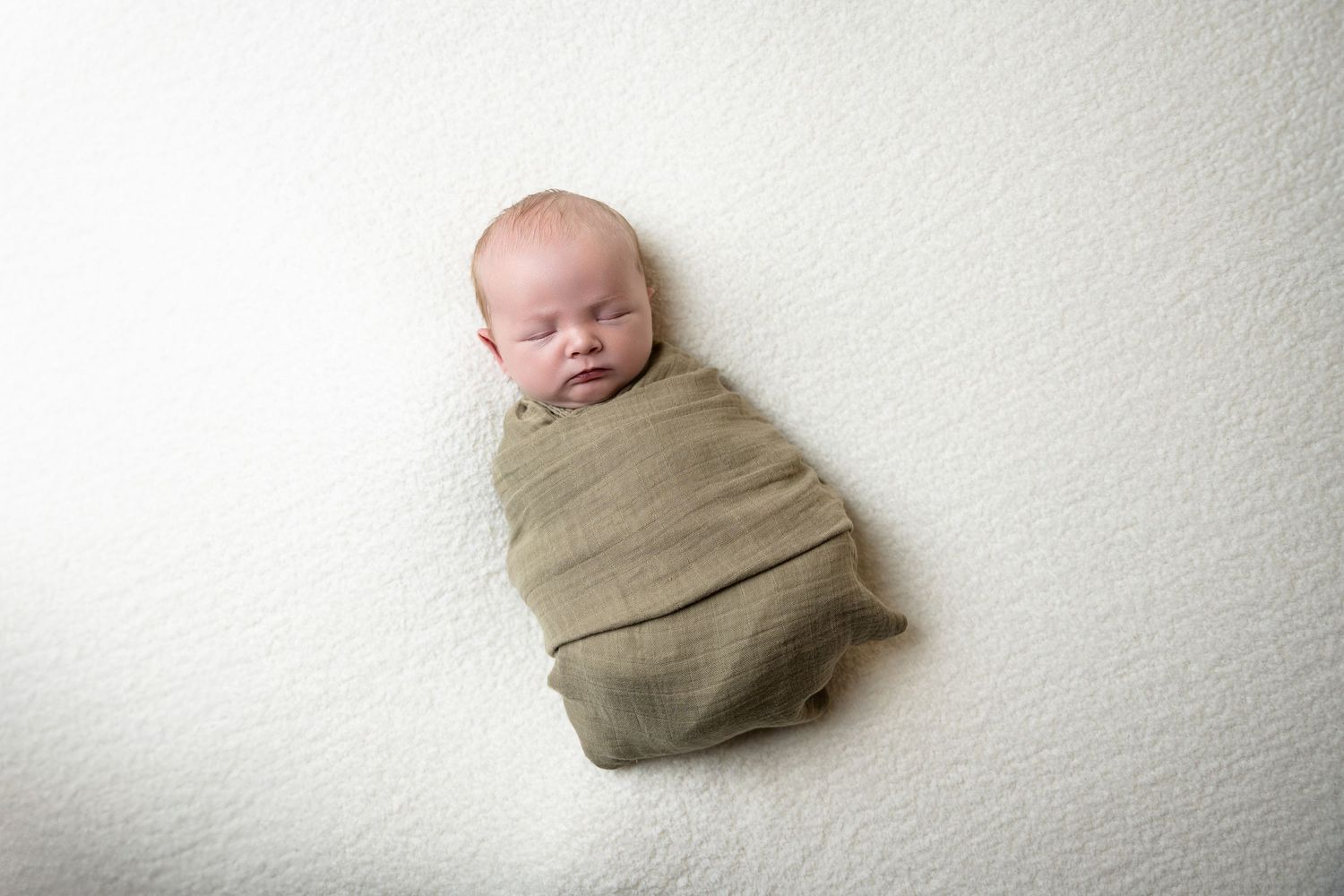 Overhead view of sleeping newborn wrapped in tan swaddle on white backdrop.
