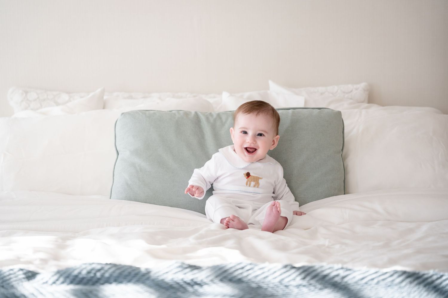 Candid lifestyle photo of baby smiling on white bedding with sage green accents.