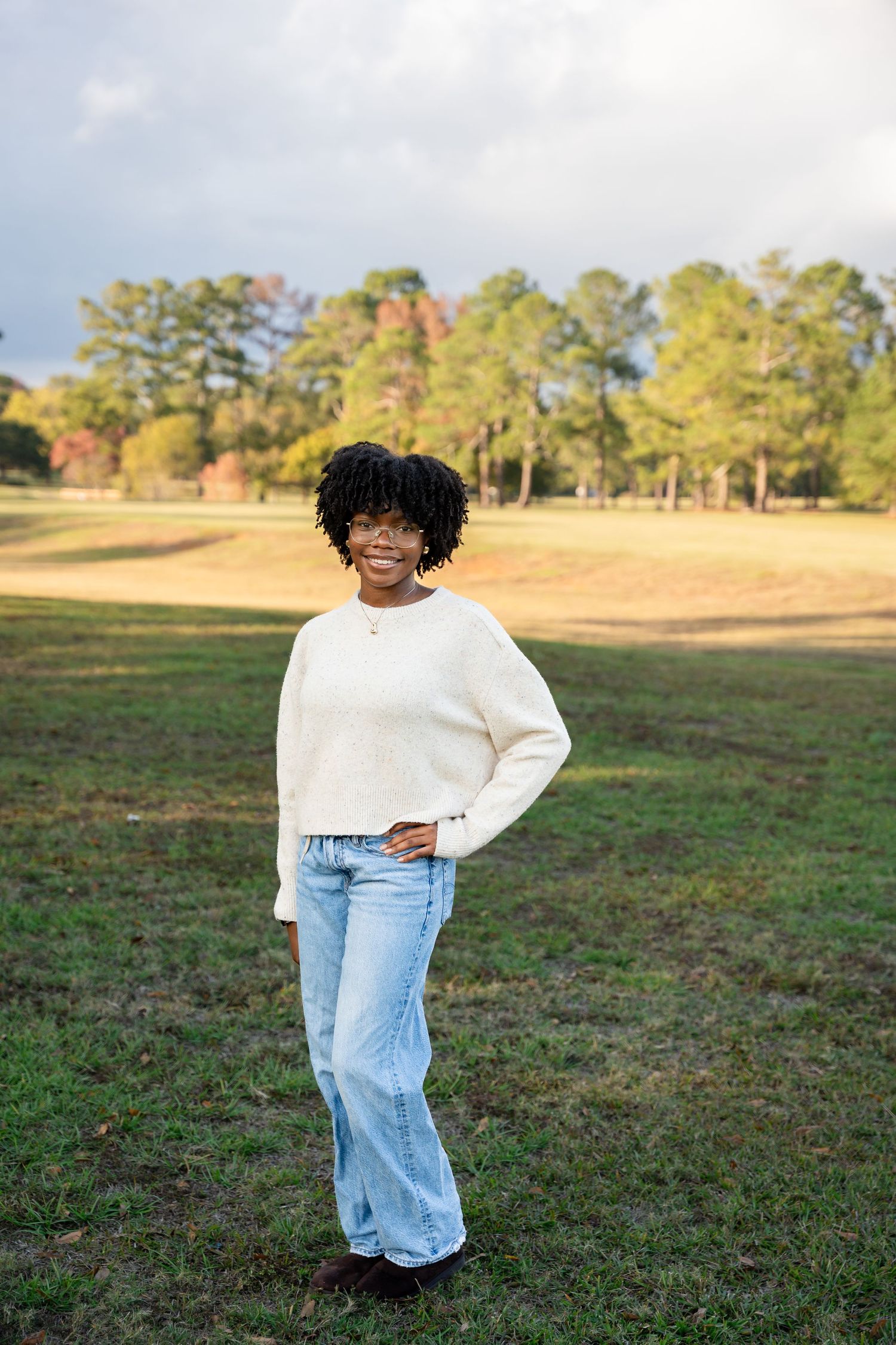 Person in white sweater and light denim poses in open field during golden hour.