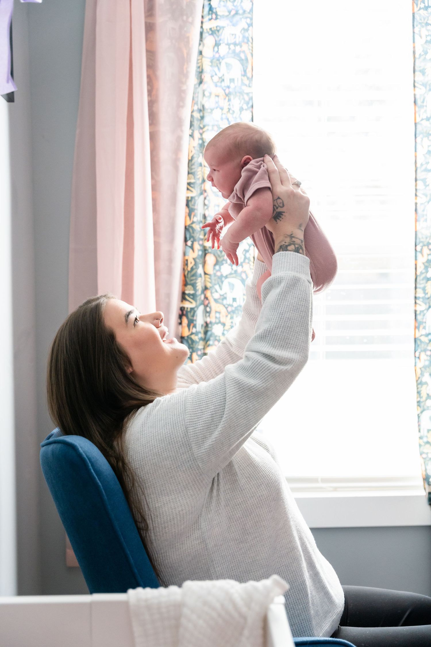 A smiling mother lifts her newborn baby in the air while sitting in a nursery chair near a bright window.