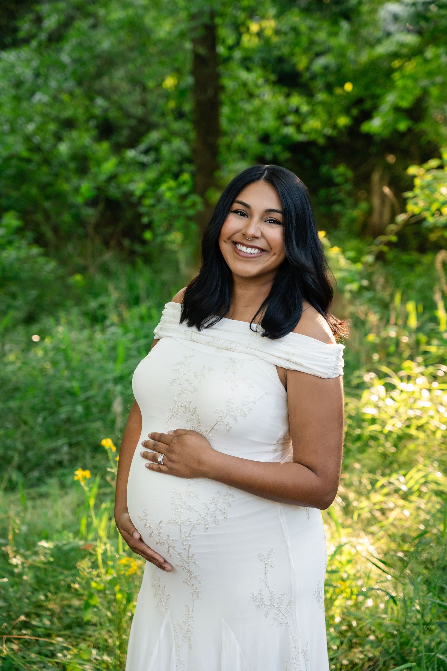 Smiling pregnant woman in white off-shoulder gown holding baby bump in a lush garden setting.