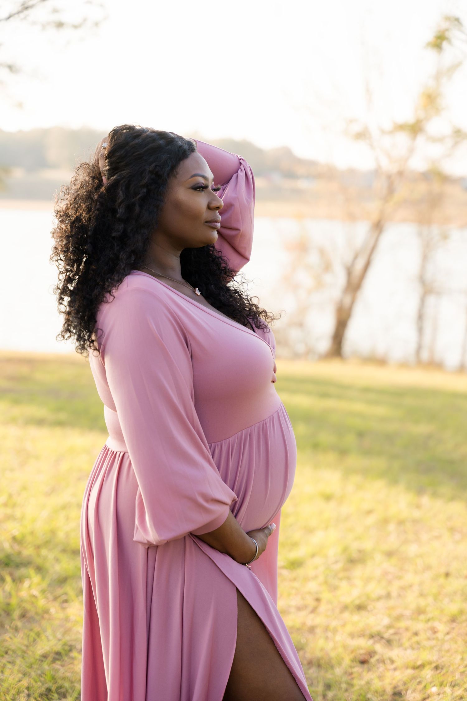 Pregnant woman in pink maxi dress poses outdoors near lake at golden hour, cradling baby bump with curly hair flowing.