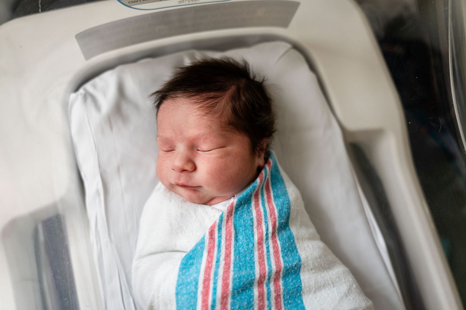 Sleeping newborn baby in hospital bassinet wearing striped hospital blanket.