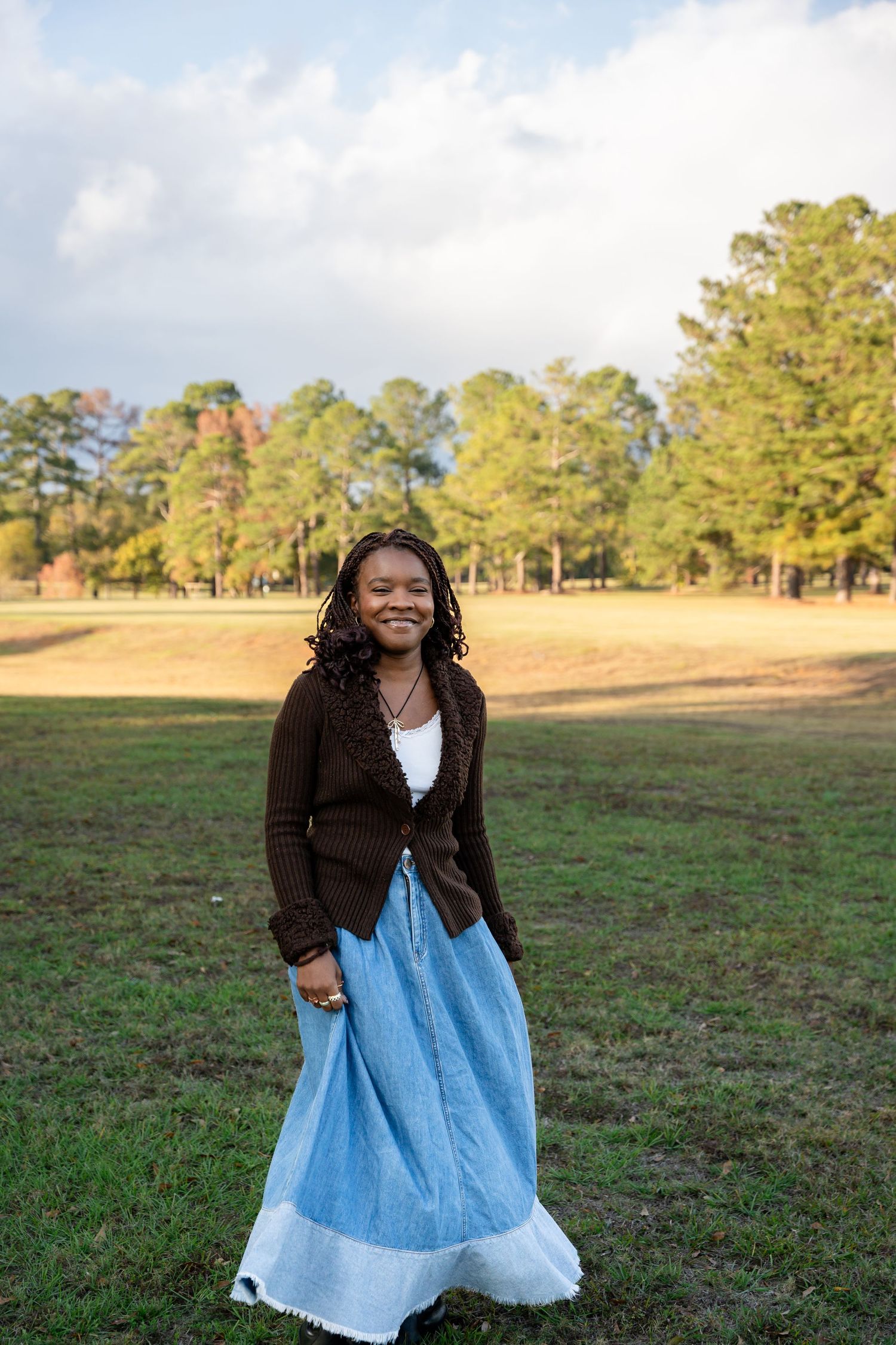 Person in blue dress and dark cardigan poses cheerfully in sunny outdoor field.