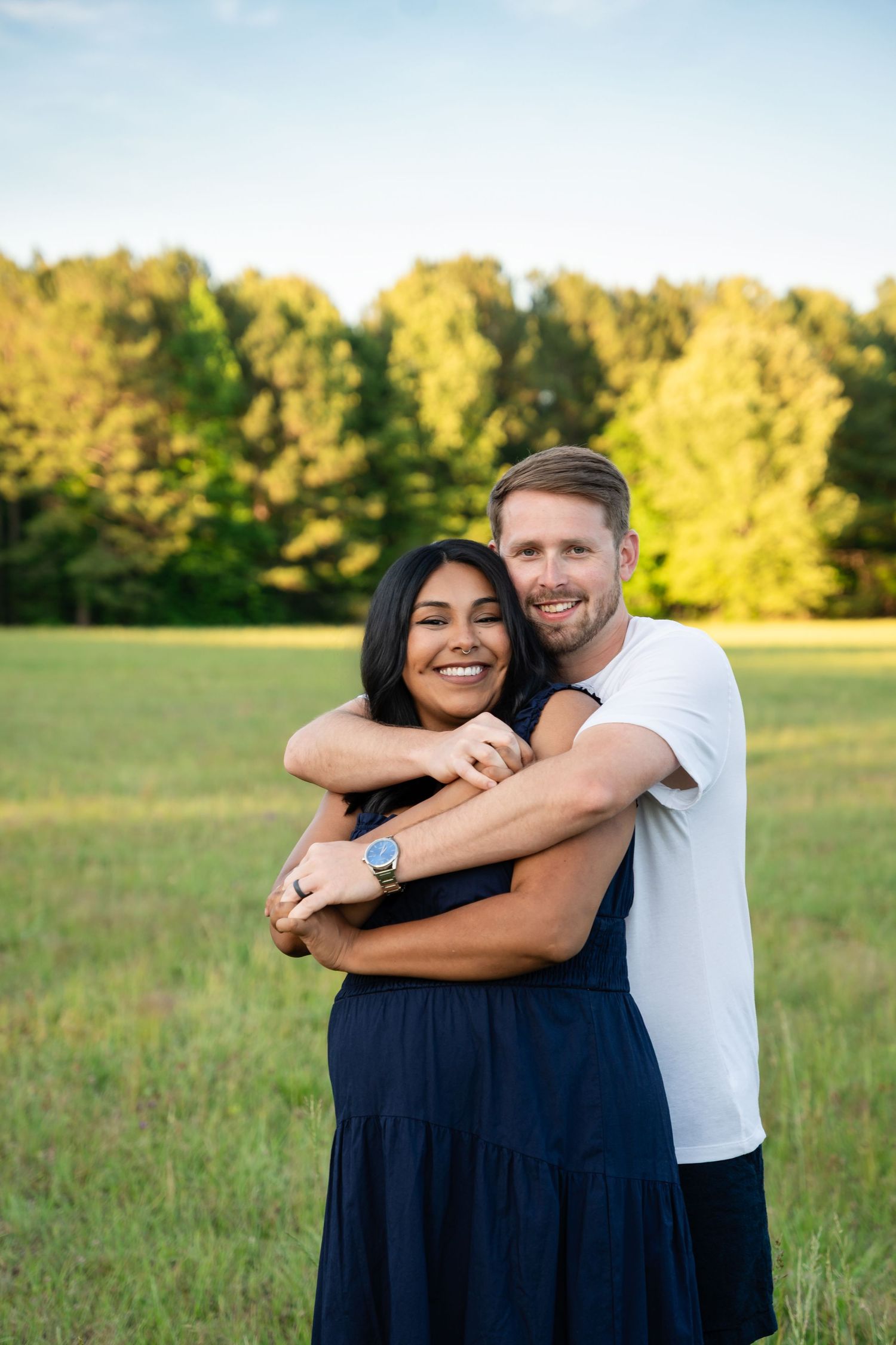 Smiling couple embraces in a green meadow at sunset as a man hugs a woman in a navy dress from behind during an outdoor photo session.