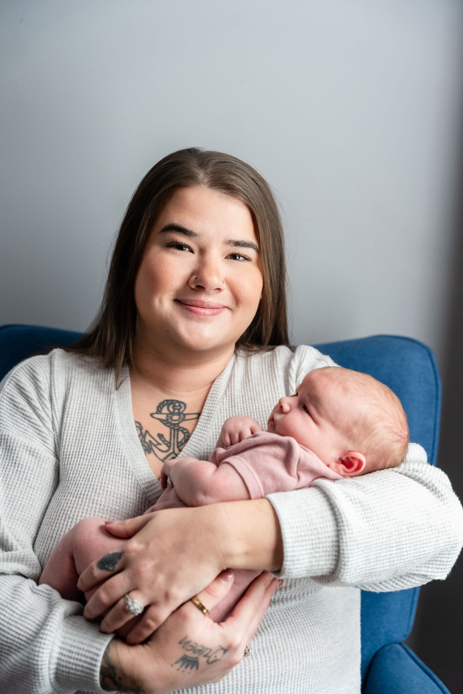 Smiling young mother with tattoos holds sleeping newborn baby in pink outfit while sitting in blue chair.