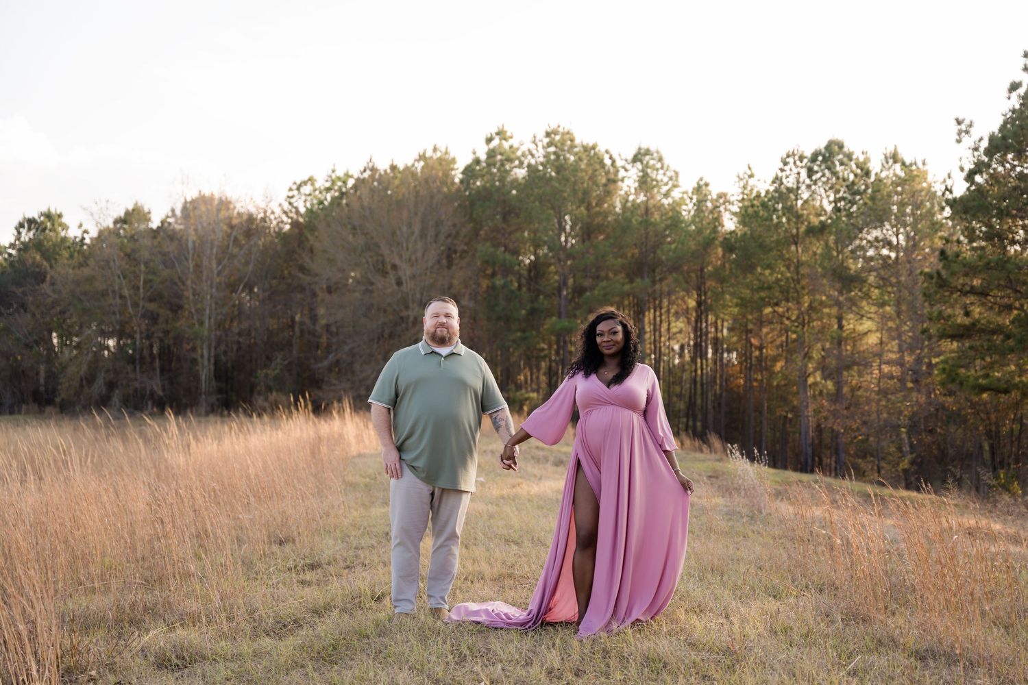 Pregnant couple holding hands in a golden field at sunset, woman in flowing pink maternity gown, trees in background.