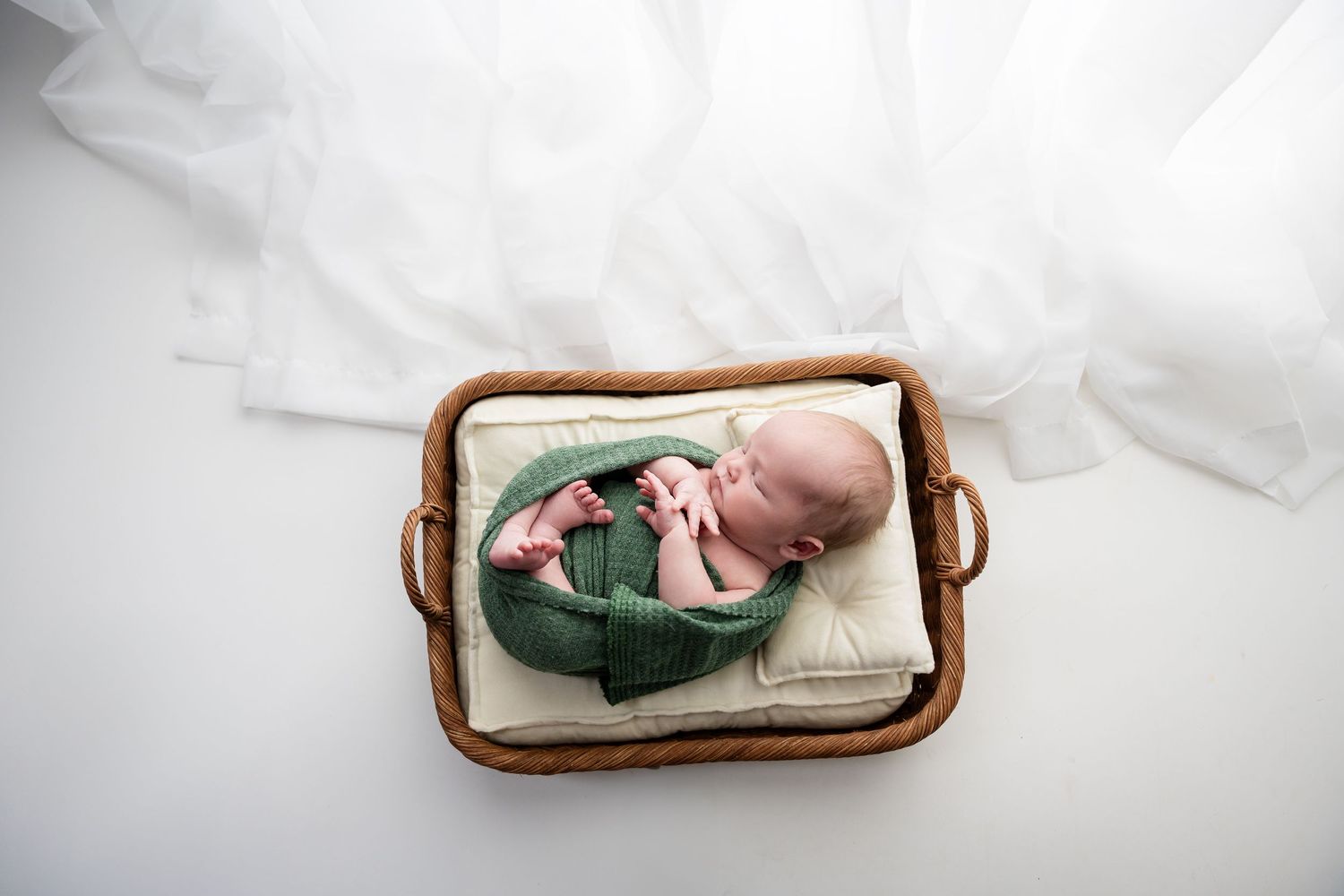 Newborn sleeping in wooden basket surrounded by white linens.