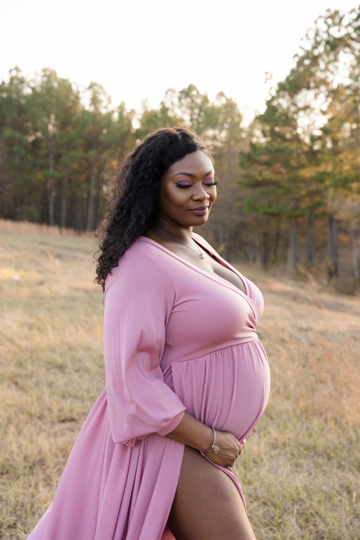 Pregnant woman in pink maternity dress cradling baby bump in sunlit outdoor field.