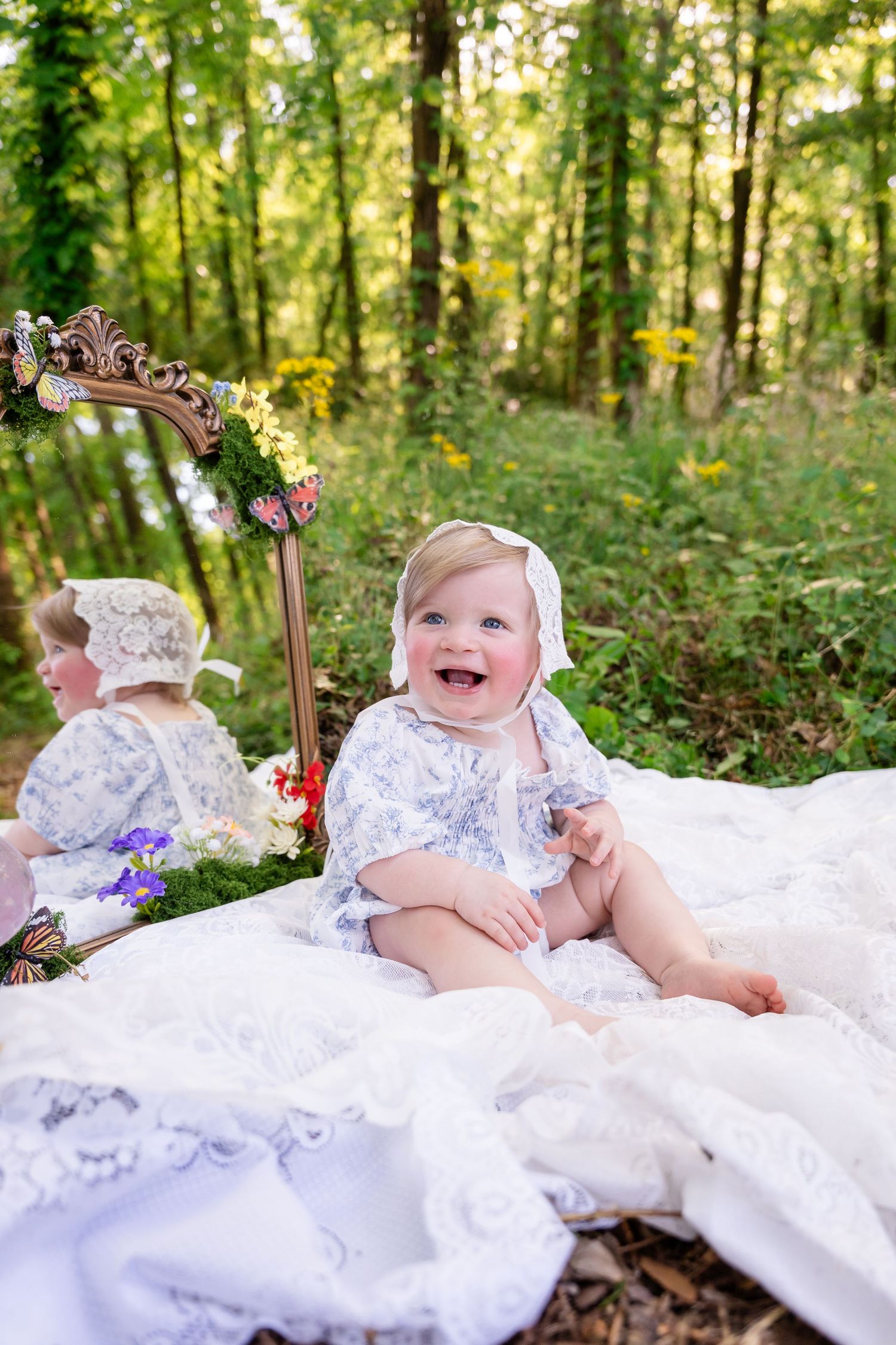 Smiling baby girl in white lace outfit and bonnet sitting on blanket outdoors in green forest setting.
