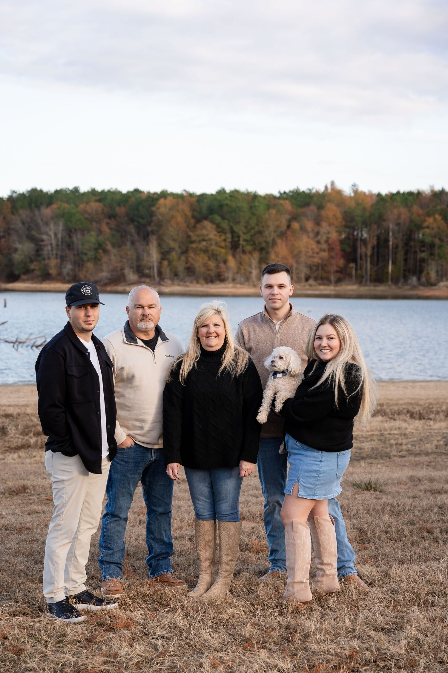 Family of five with a small white dog posing outdoors in a grassy field by a lake, with autumn trees in the background.