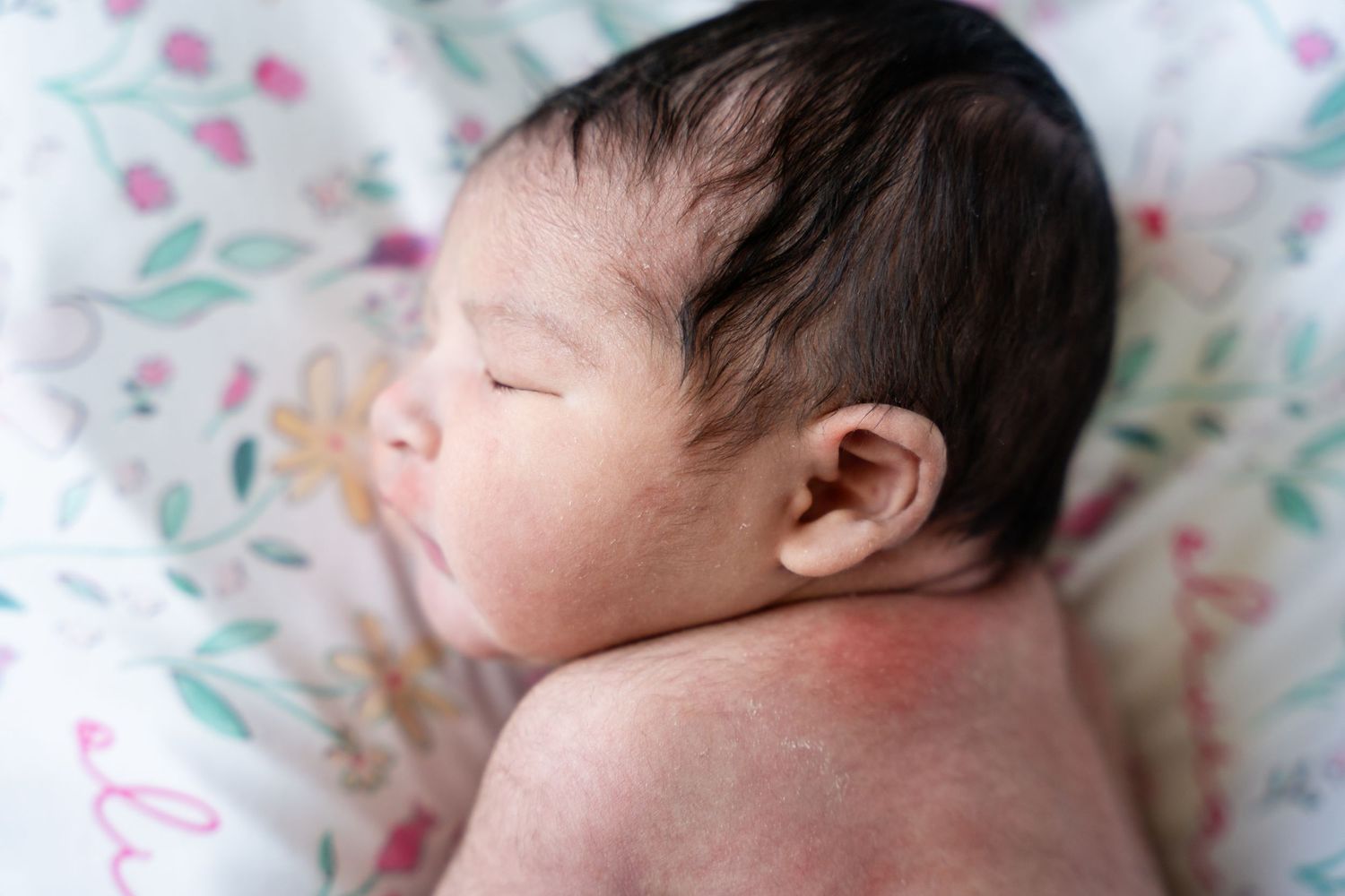 Sleeping newborn baby with dark hair lying on a soft floral-patterned blanket, shown in profile with bare shoulders.