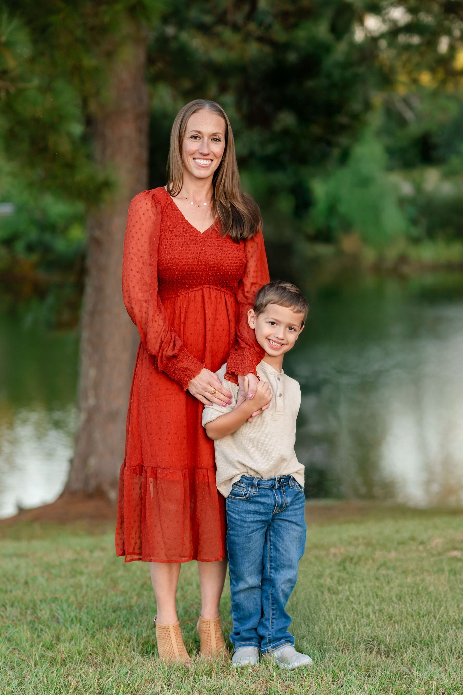 A smiling woman in a red dress poses with a young boy in jeans outdoors near a pond.