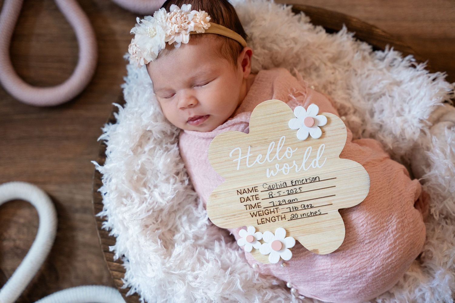 Newborn baby girl sleeping in a basket with a floral headband and a Hello World wooden birth announcement sign.