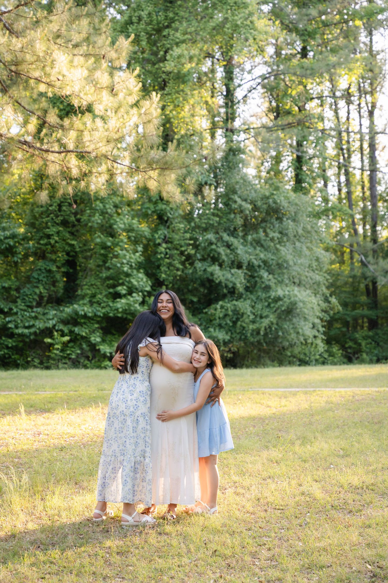 Pregnant woman in white dress embraced by two daughters in sunny green park during maternity photo session.