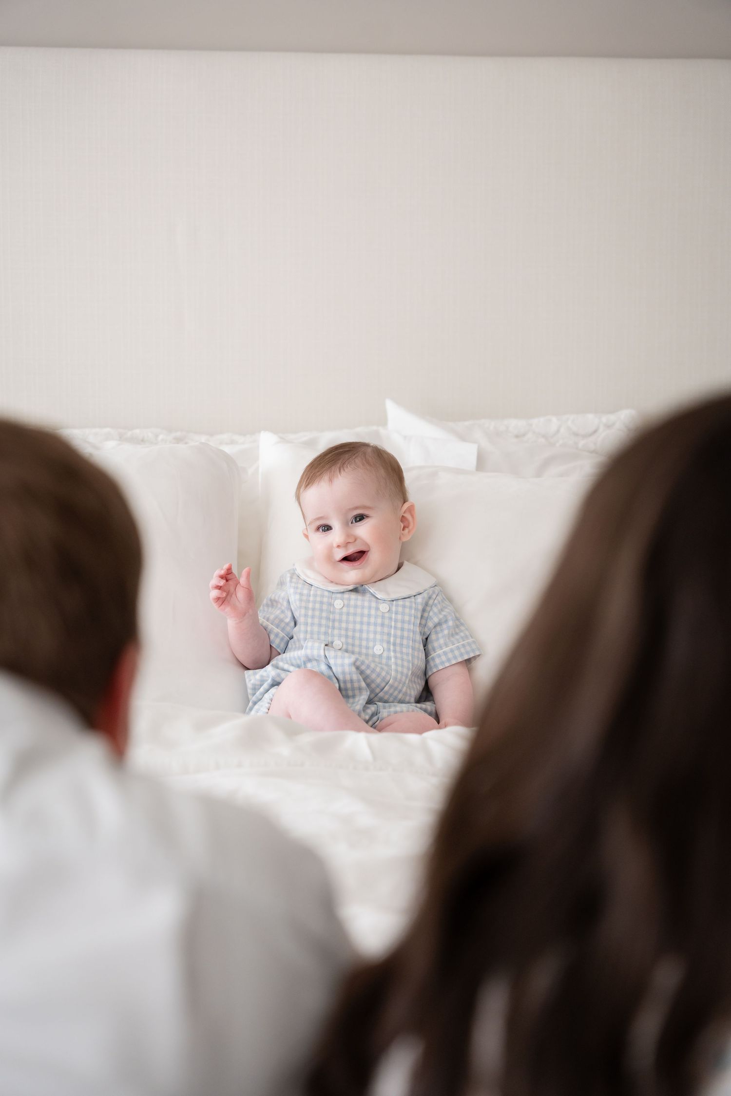 A happy baby wearing a blue plaid outfit sits on a white surface while others watch.