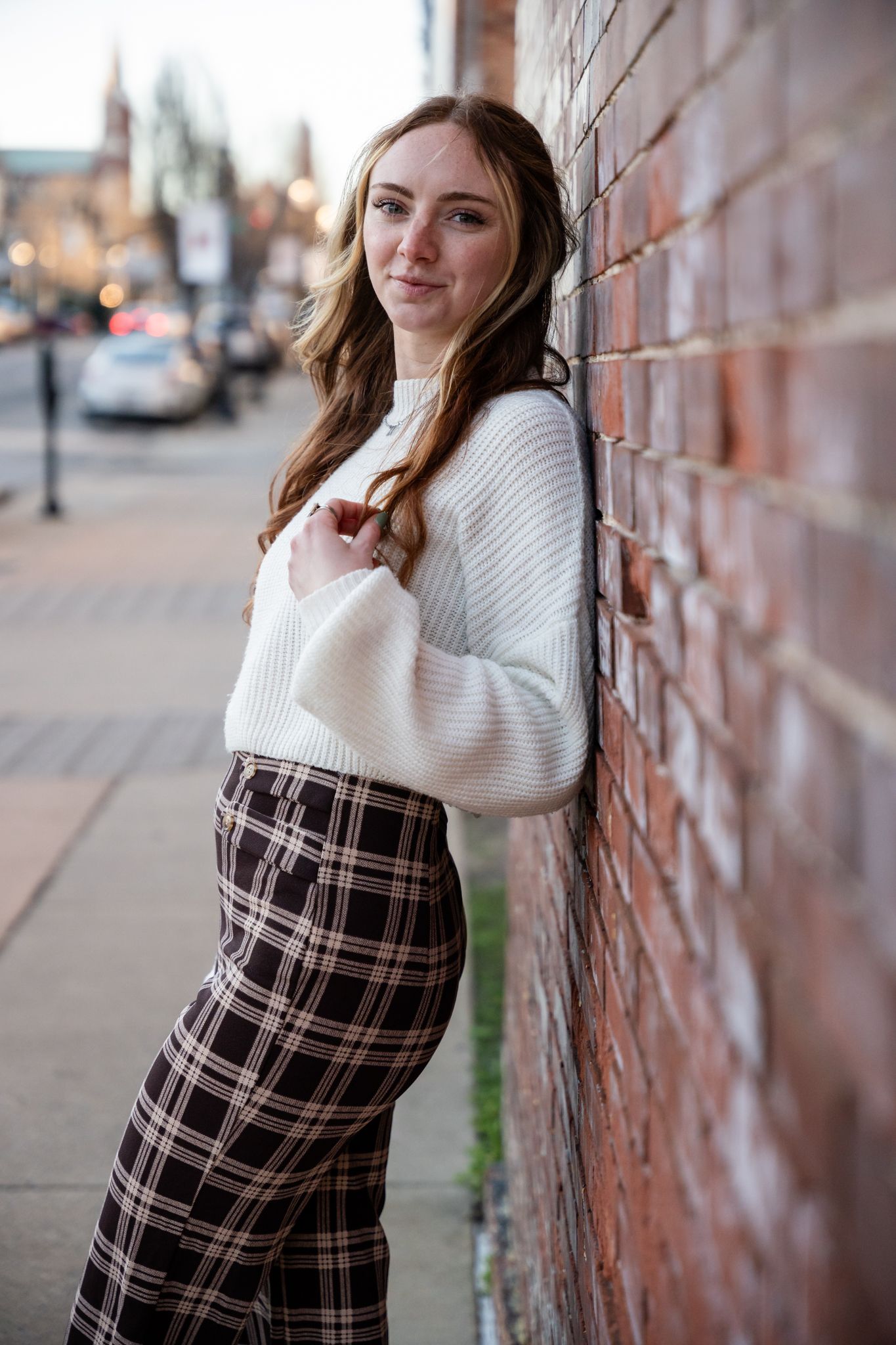 Urban fashion portraits against brick wall wearing white knit sweater and plaid pants.