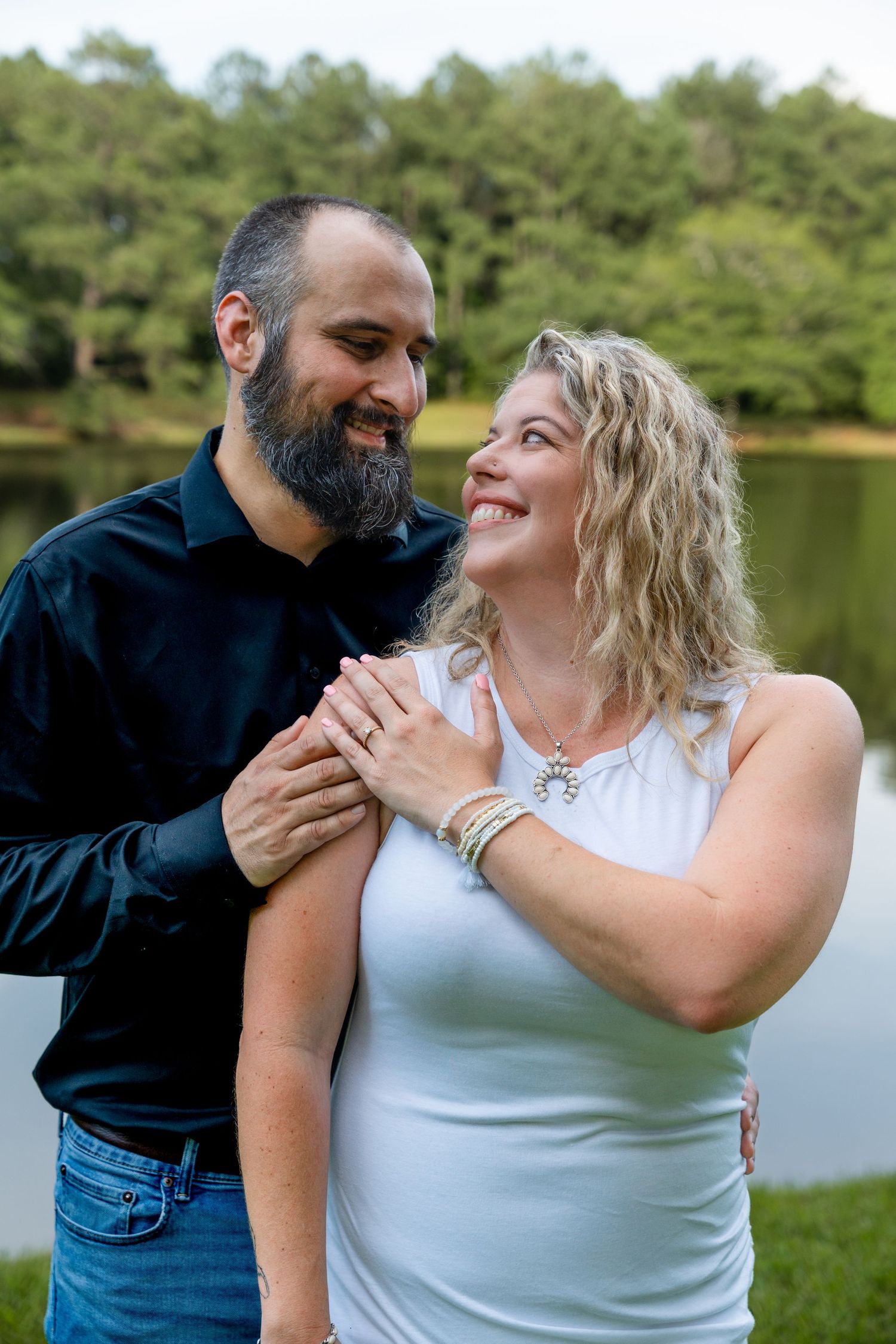 Couple sharing intimate moment by lakeside with trees in background.