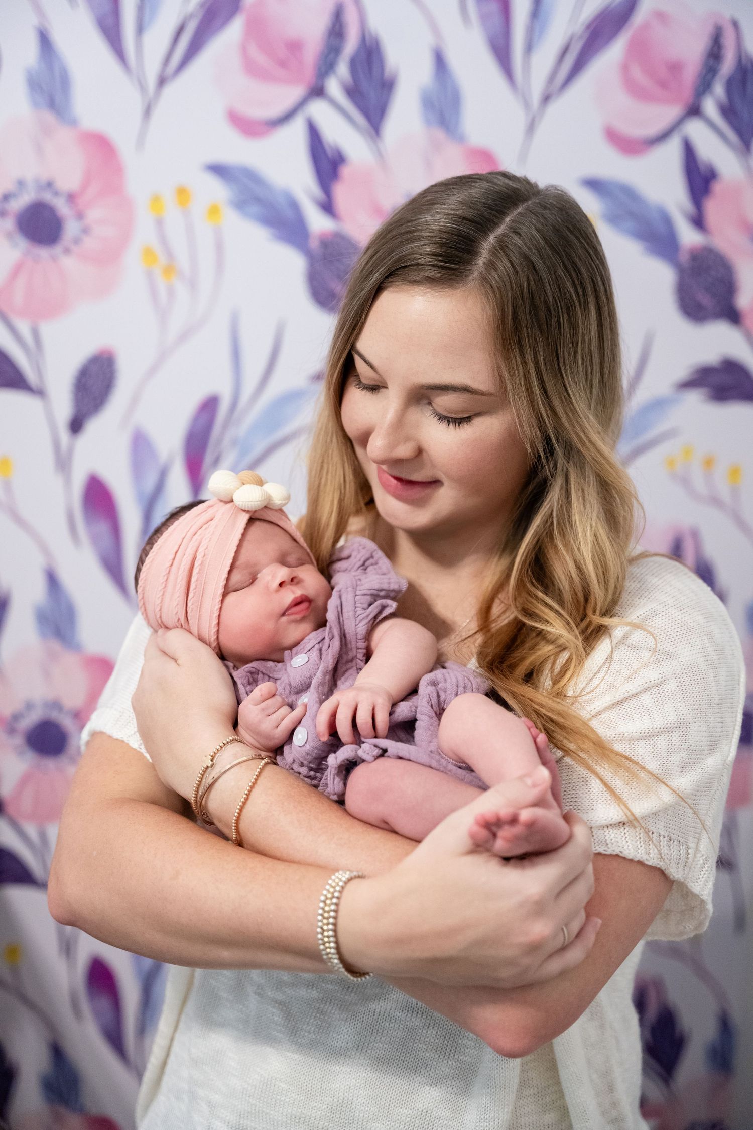 Young mother smiling while gently cradling her sleeping newborn baby wrapped in purple against a floral wallpaper backdrop.
