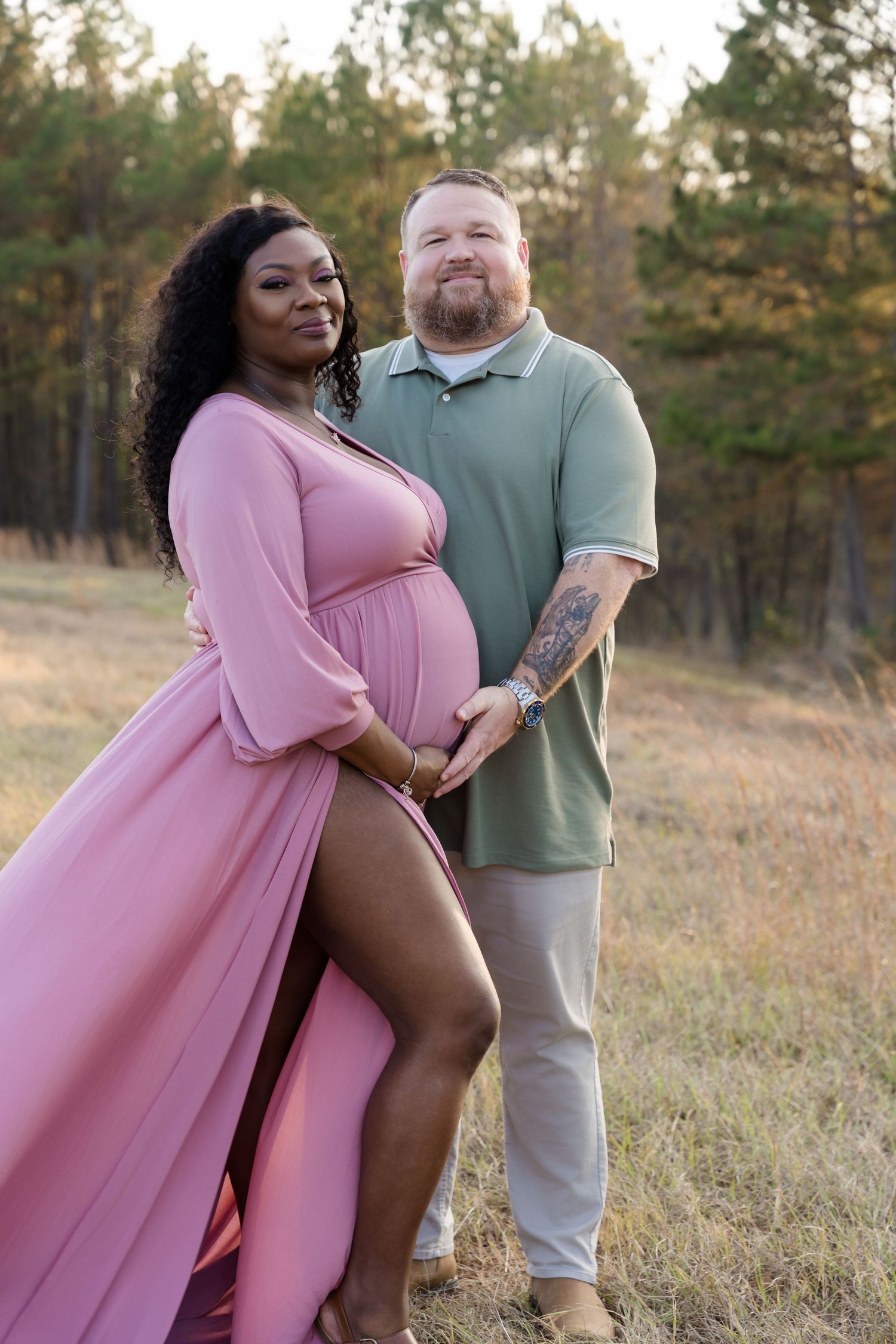 Interracial couple posing together in field, woman wearing pink maternity gown at sunset.