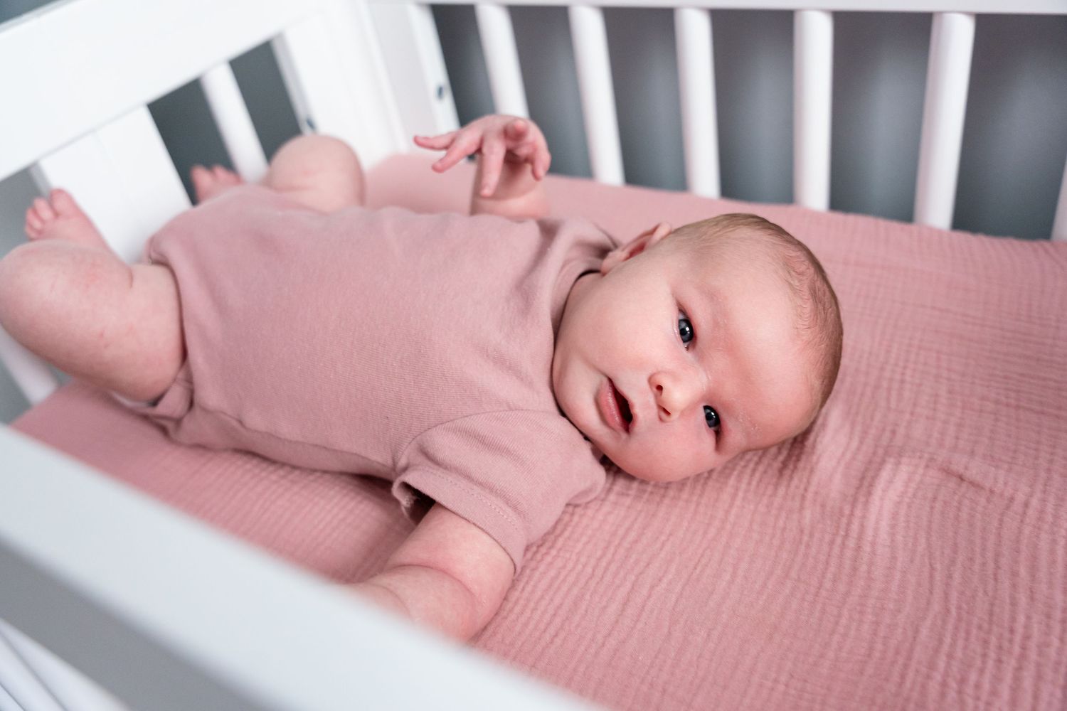 Newborn baby lying awake in a white crib on a pink blanket wearing a pink onesie.