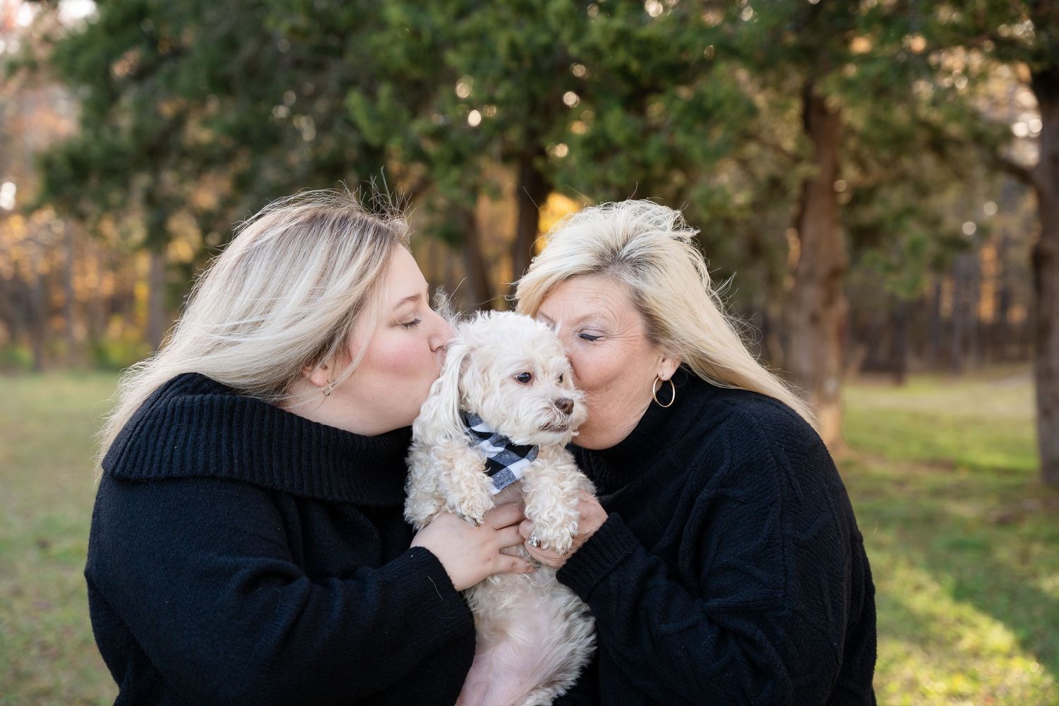 Two women with blonde hair in black sweaters lovingly kiss and hold a fluffy white puppy wearing a bandana in a sunny outdoor park.