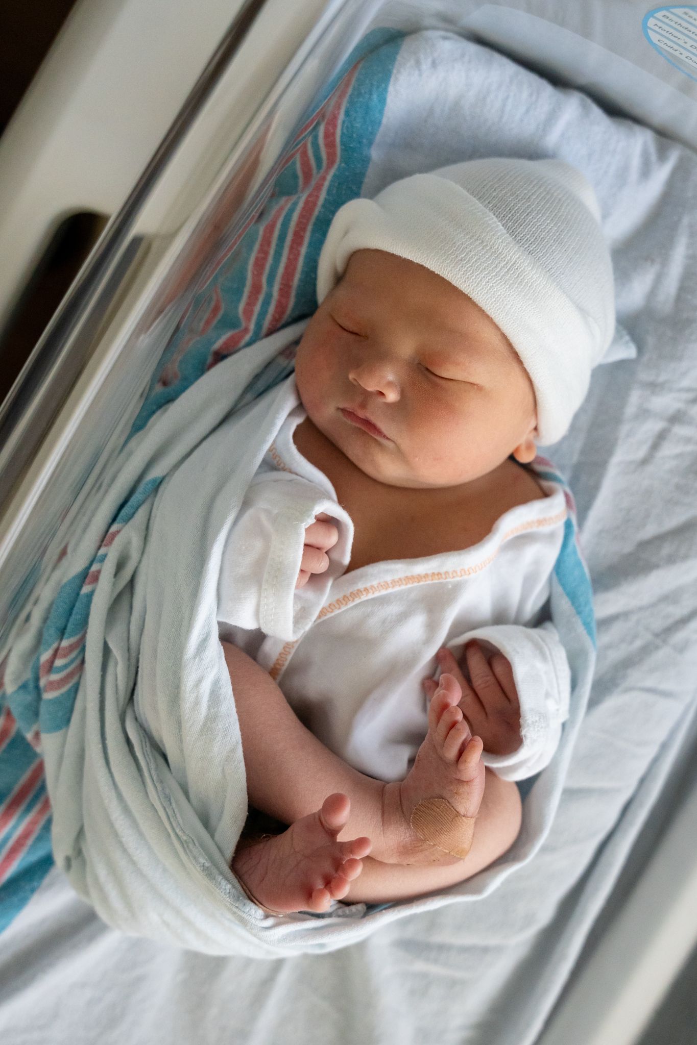 Newborn baby wearing white cap and swaddle blanket resting peacefully.