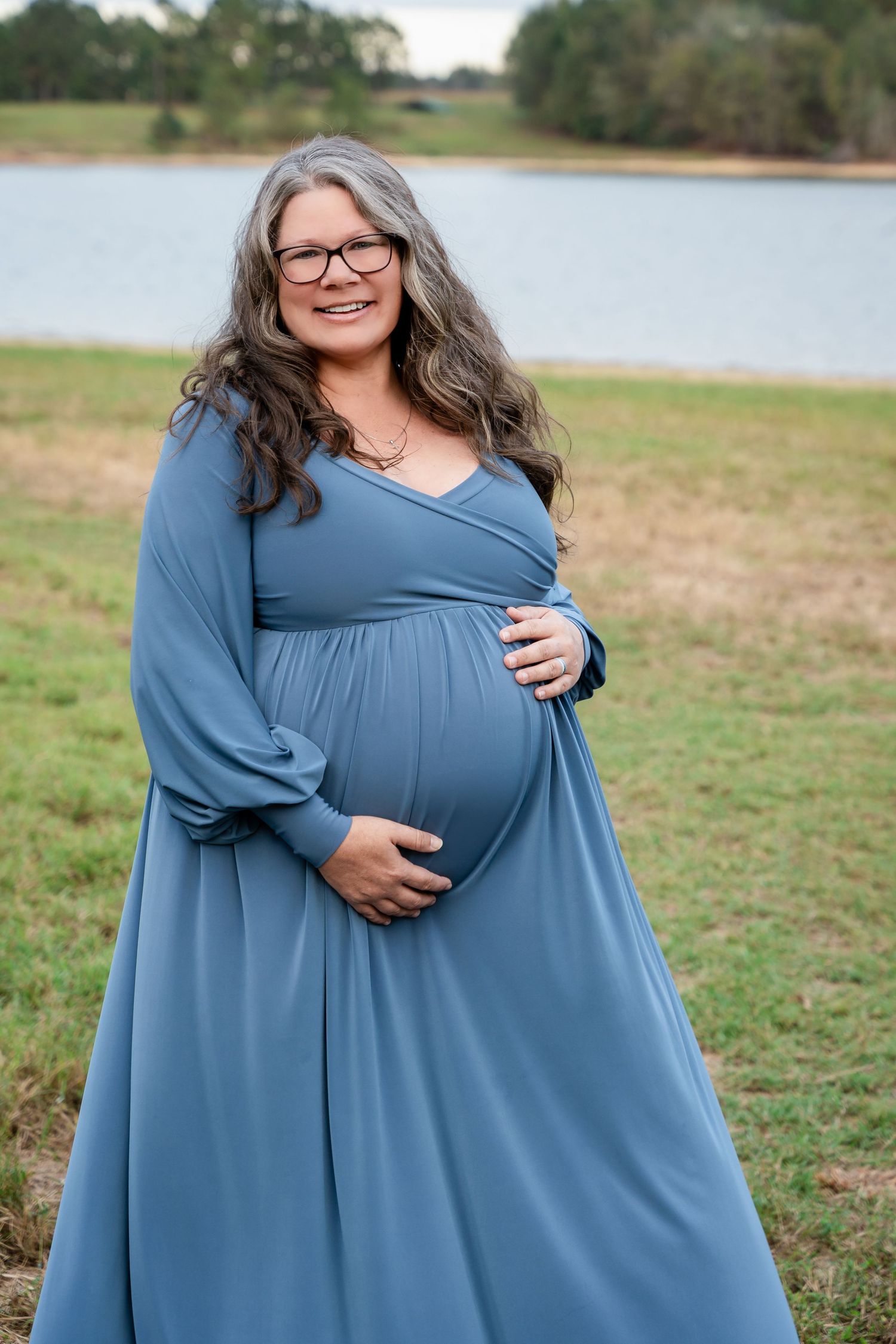 Woman in flowing blue dress poses for maternity photos in outdoor field setting.