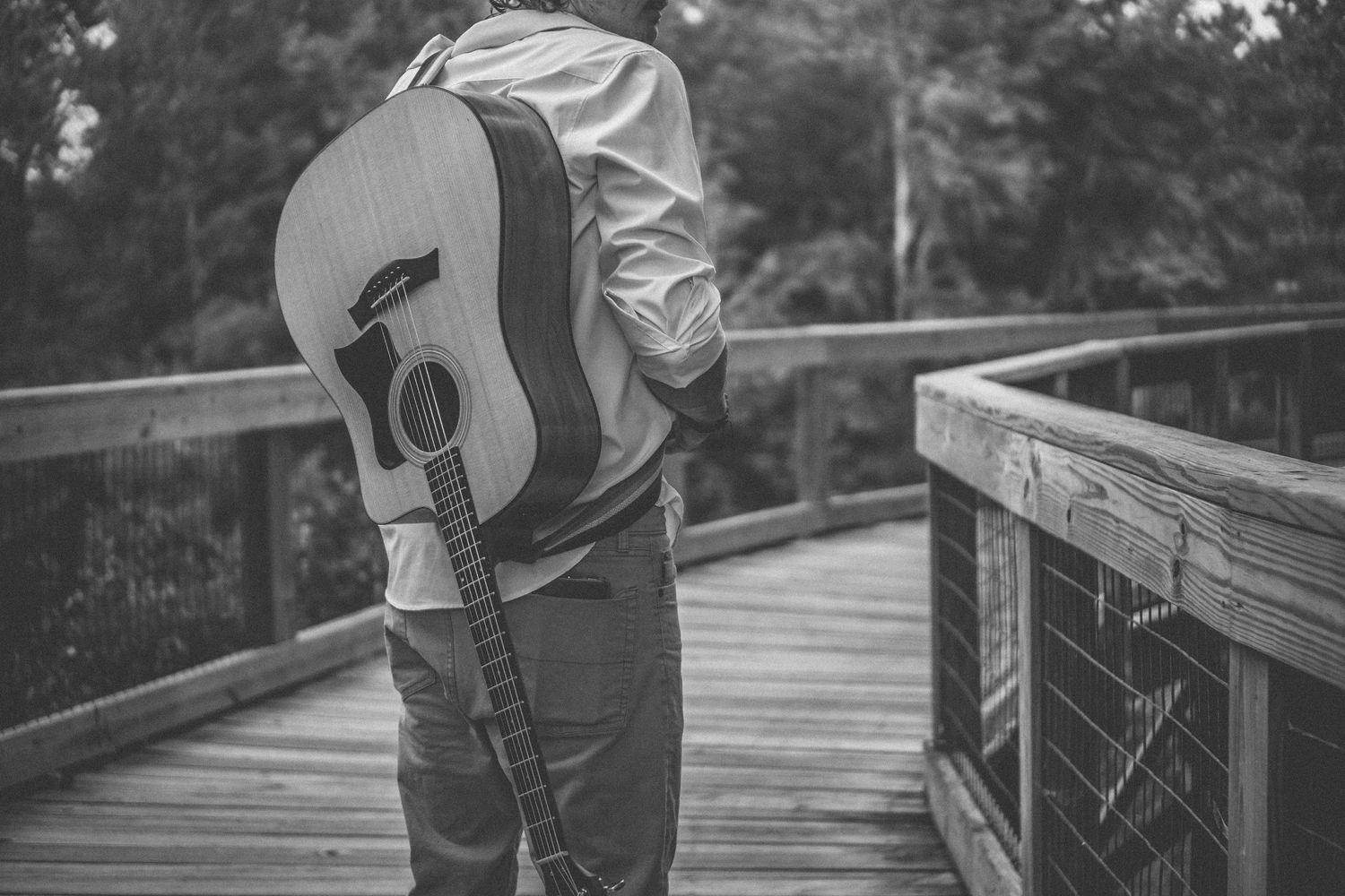 Black and white image of a musician holding an acoustic guitar while standing on a wooden boardwalk.