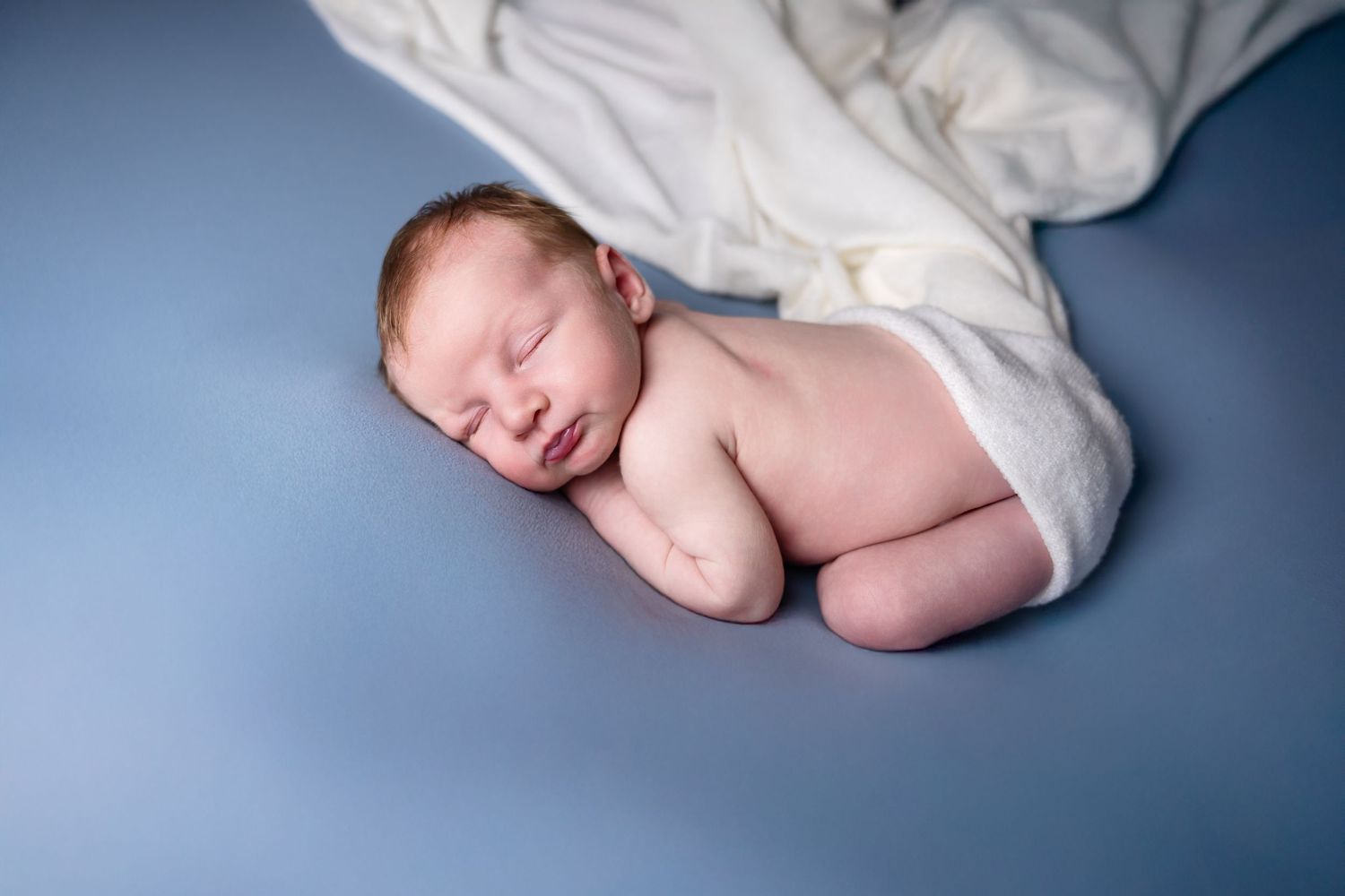 Minimalist newborn photography with baby wrapped in white cloth against dark blue background.