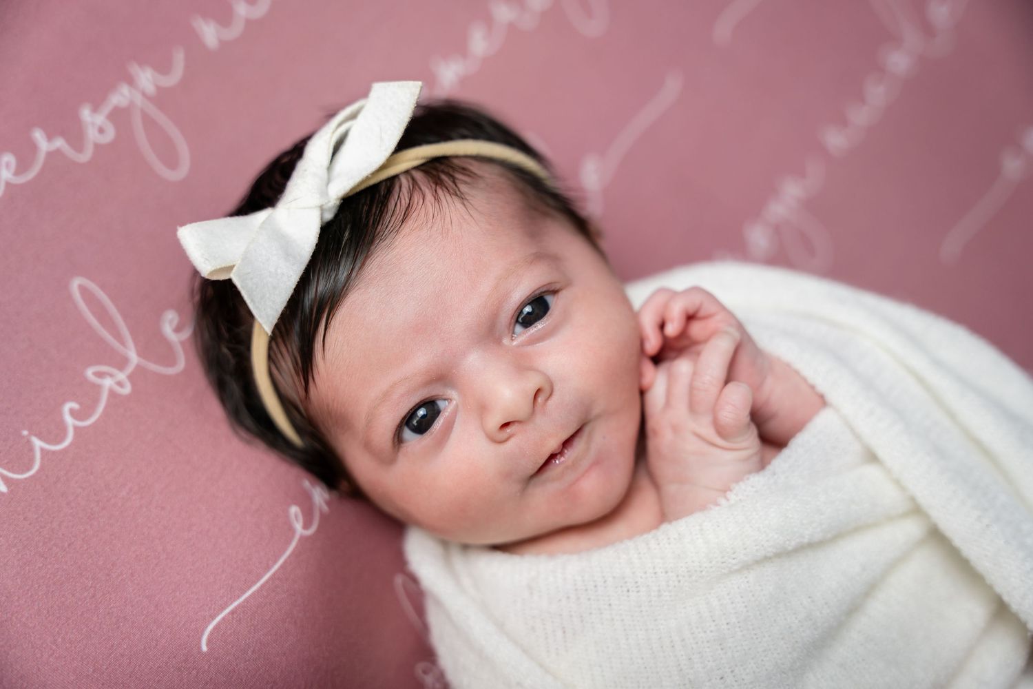 A newborn baby wearing a white bow headband lies on pink fabric while making various sweet expressions.
