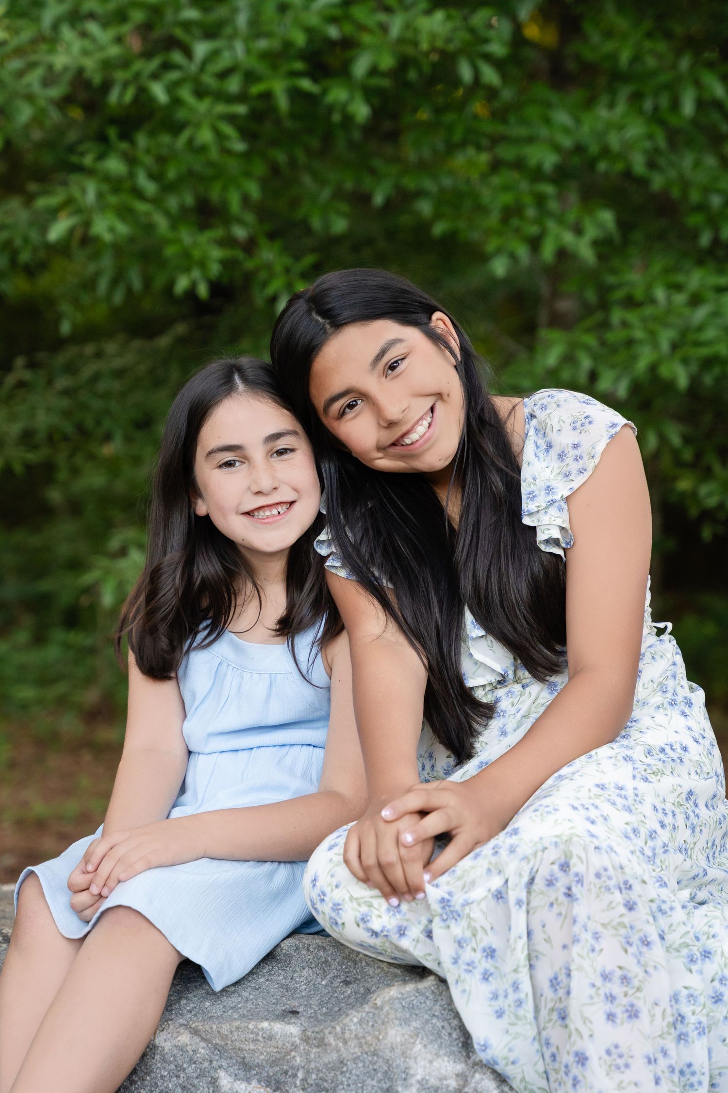 Two smiling sisters in blue dresses sit on a rock outdoors, surrounded by lush green trees.